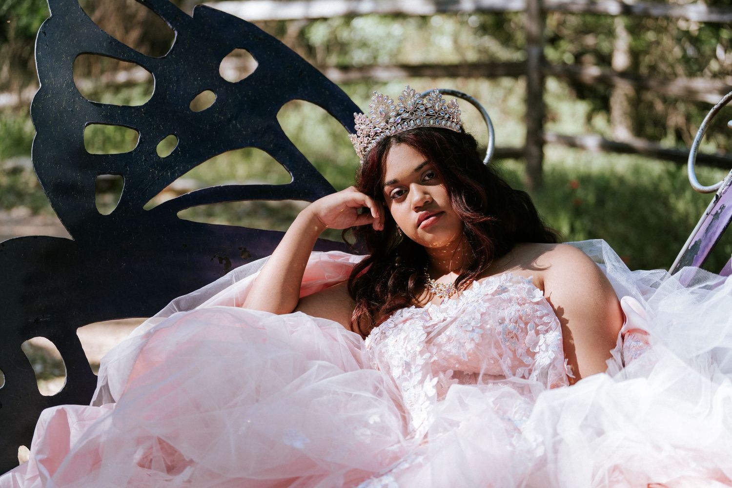 Young woman in pink ball gown and tiara reclines on decorative butterfly bench outdoors in elegant quinceañera portrait.