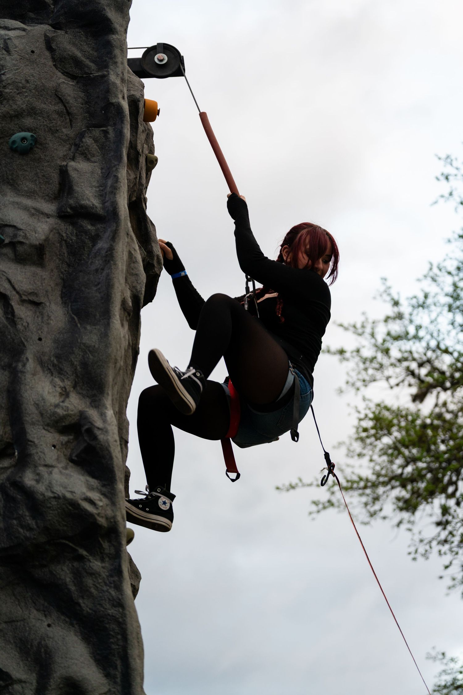 A red-haired climber scales an outdoor rock wall, suspended by safety harness and rope against a cloudy sky.