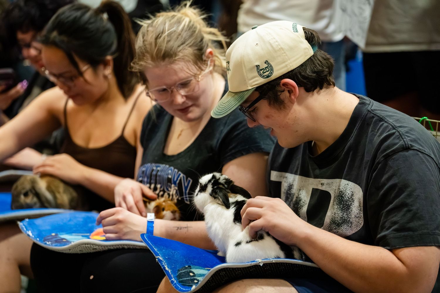 Two students read a book together while holding a small rabbit at an indoor animal therapy event.