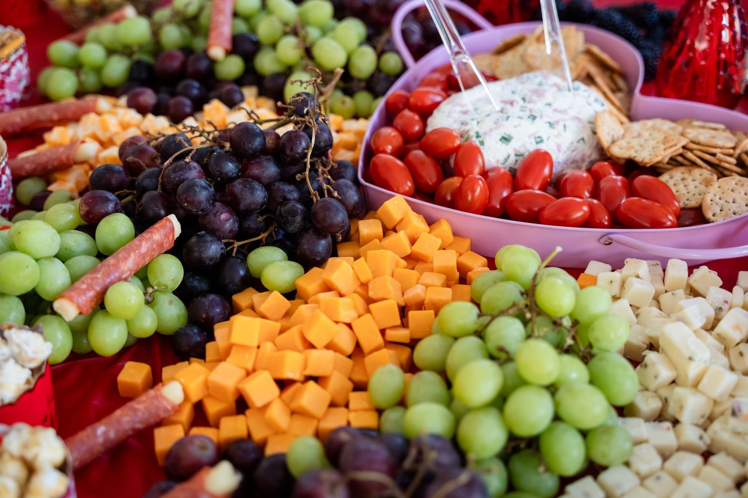 Colorful charcuterie board with green and purple grapes, cheddar cubes, cherry tomatoes, crackers, and creamy dip.