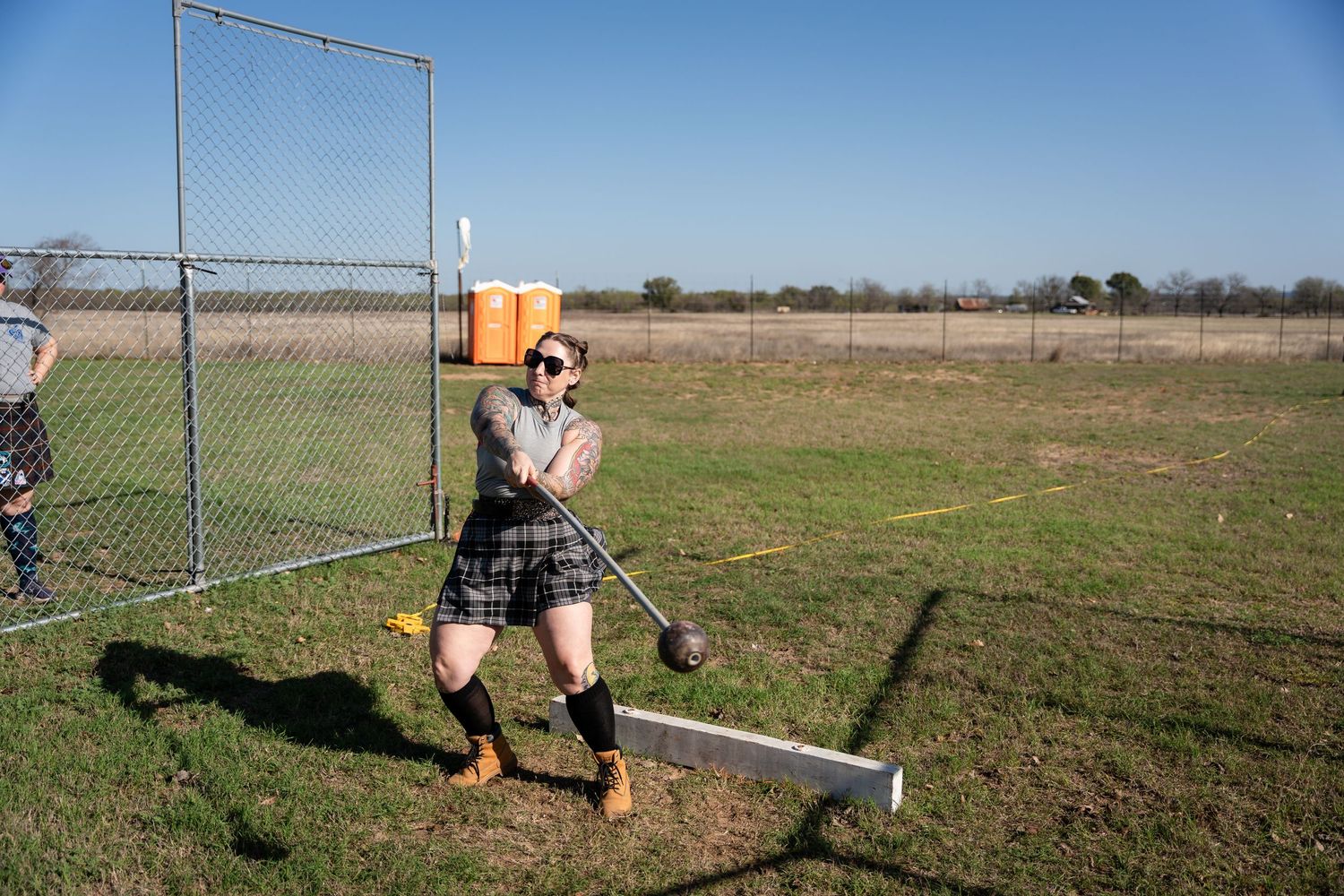 Man in plaid kilt throwing hammer at Highland Games competition on green field.
