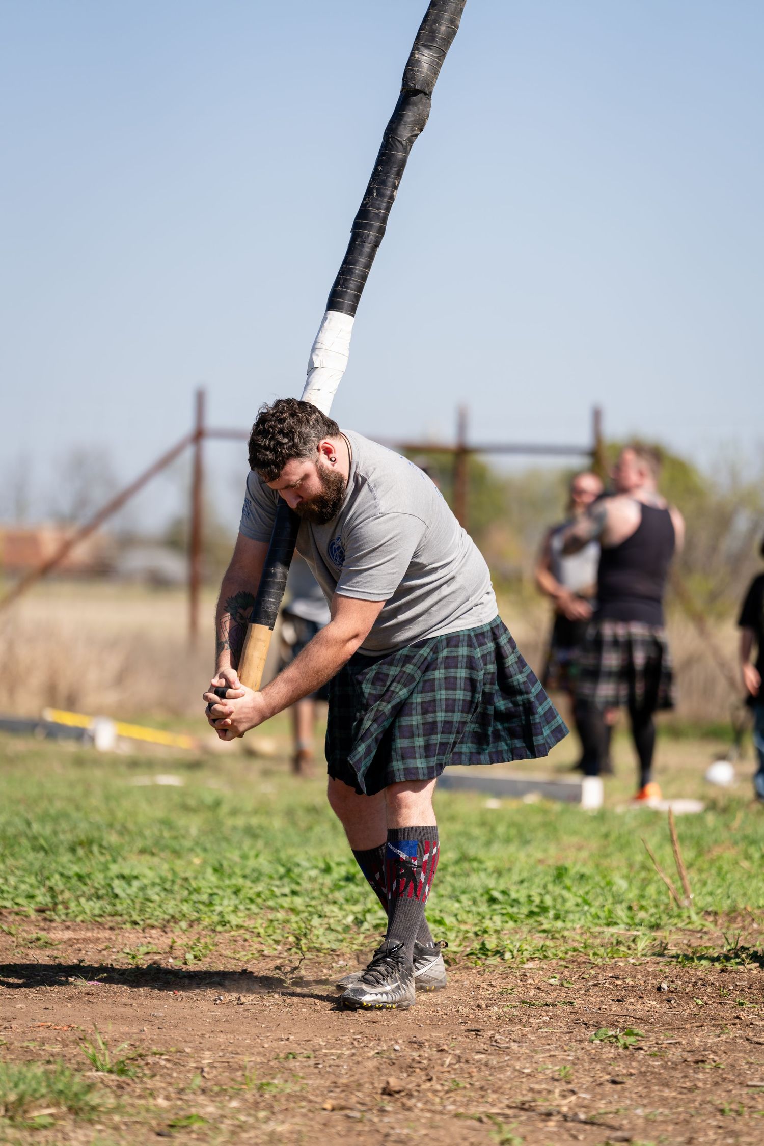 Man in tartan kilt tossing caber at Highland Games competition outdoors on sunny day.