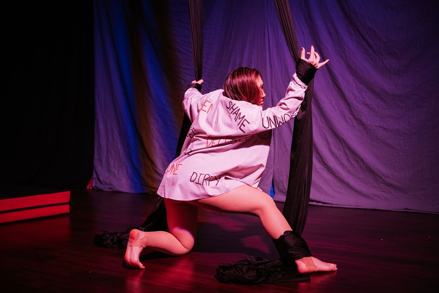 Aerial silk performer in white robe kneeling on stage with dramatic red and blue lighting.