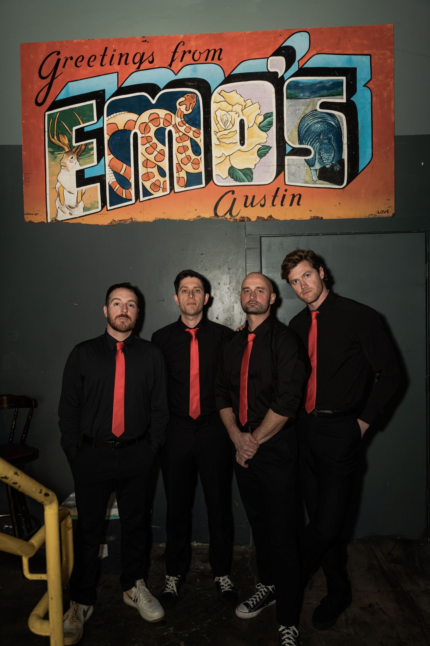 Four men in black shirts and red ties pose at Emo's Austin venue beneath a colorful Greetings from Emo's mural.