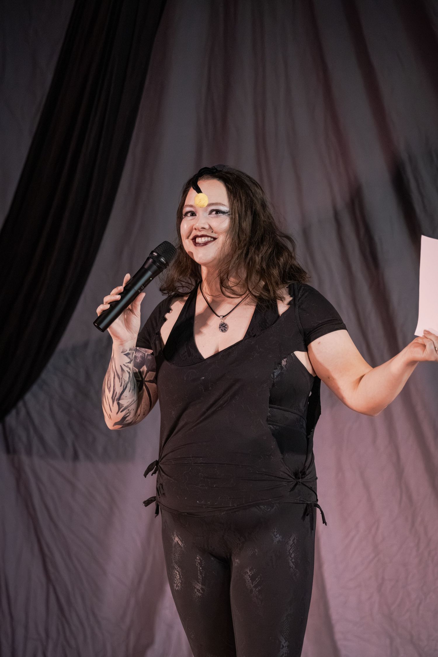 Woman with tattoos holding microphone and papers, performing on stage in black outfit with dramatic backdrop.