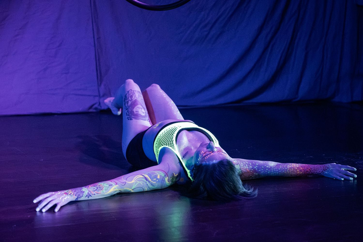 Performer with UV body paint glowing under black light, lying on stage floor in artistic pose.
