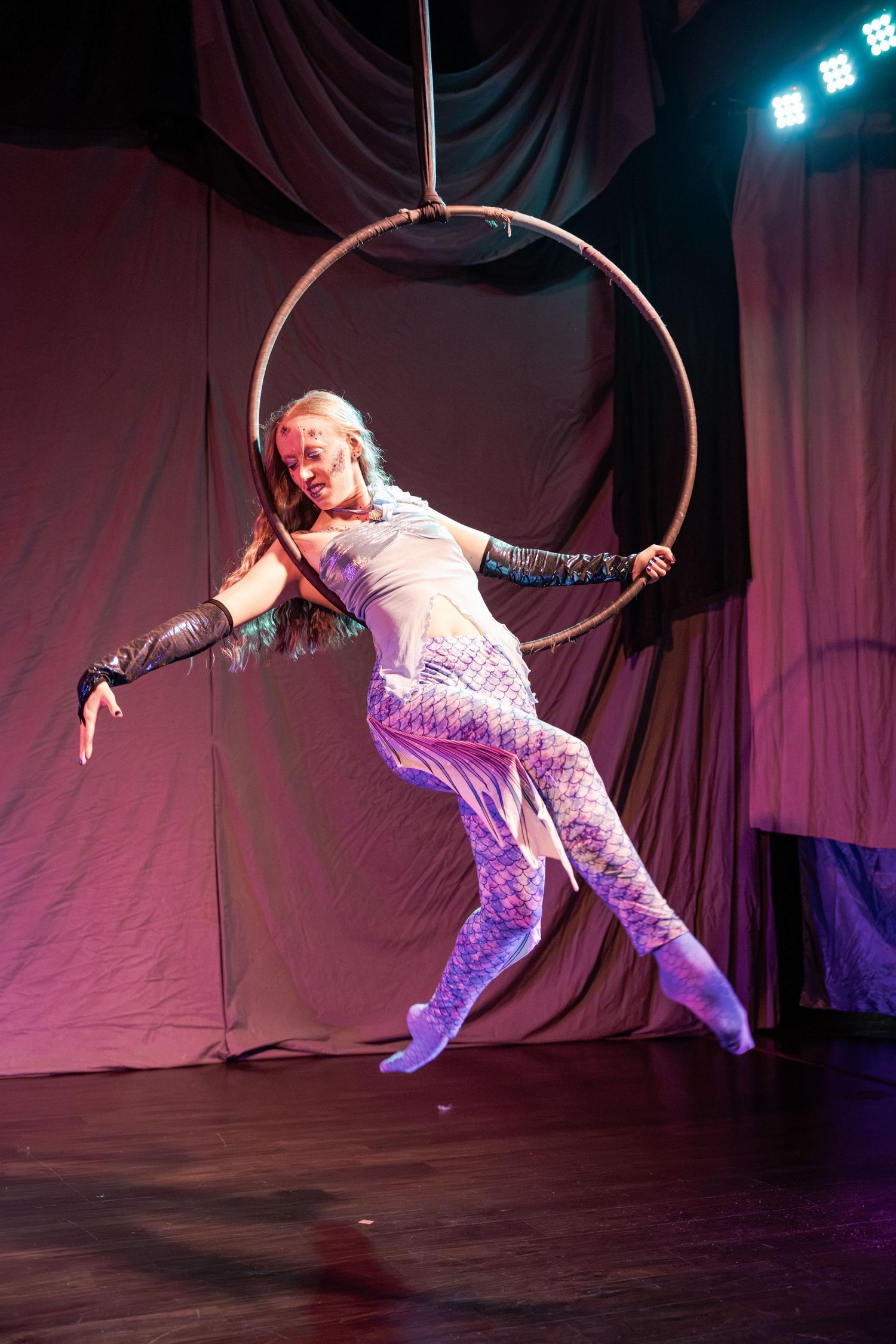 Aerial acrobat in mermaid costume performs on a lyra hoop under stage lights at a circus performance.