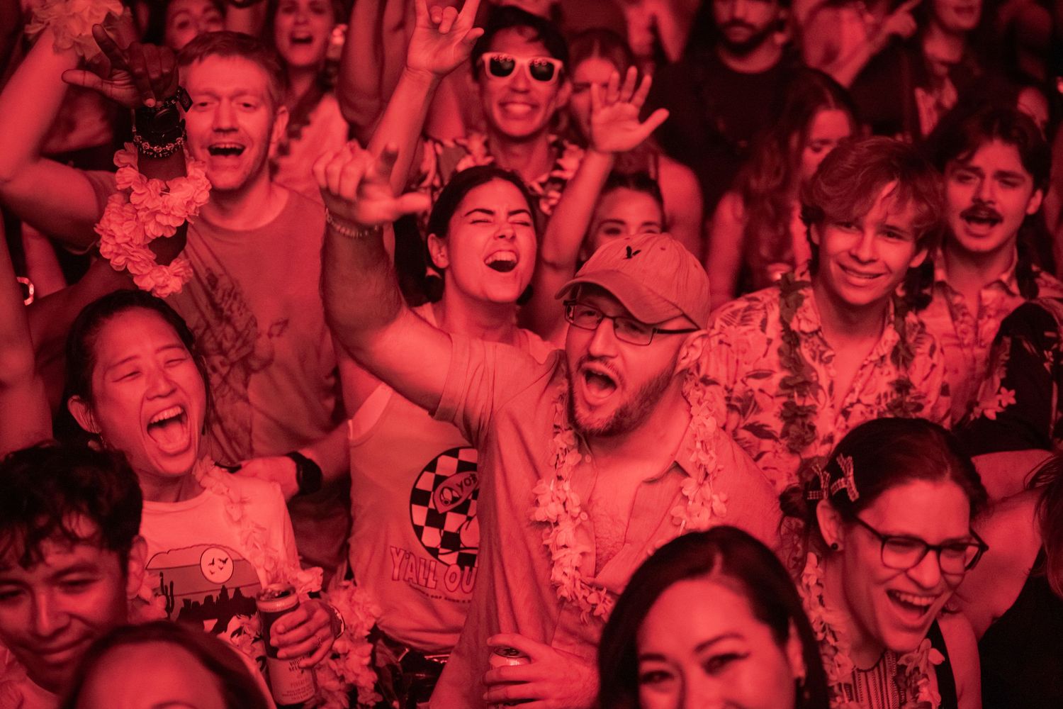 Excited crowd of diverse concert-goers cheering under red stage lighting at a live music festival.