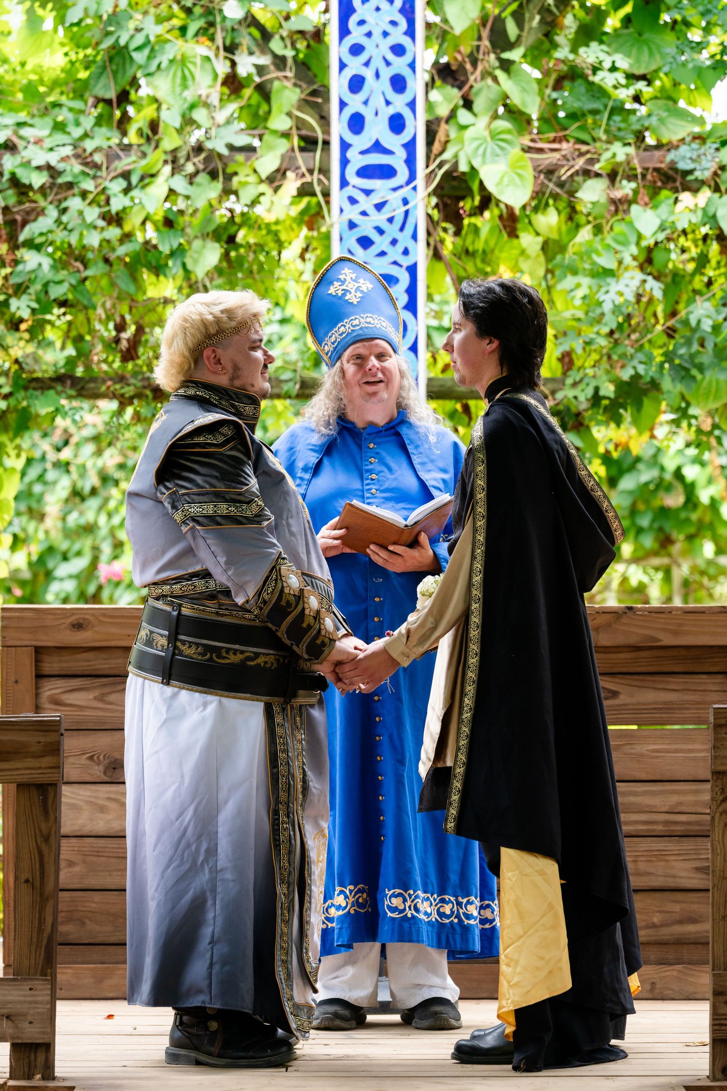 Three figures in ceremonial robes interact during an outdoor ceremony under green foliage.