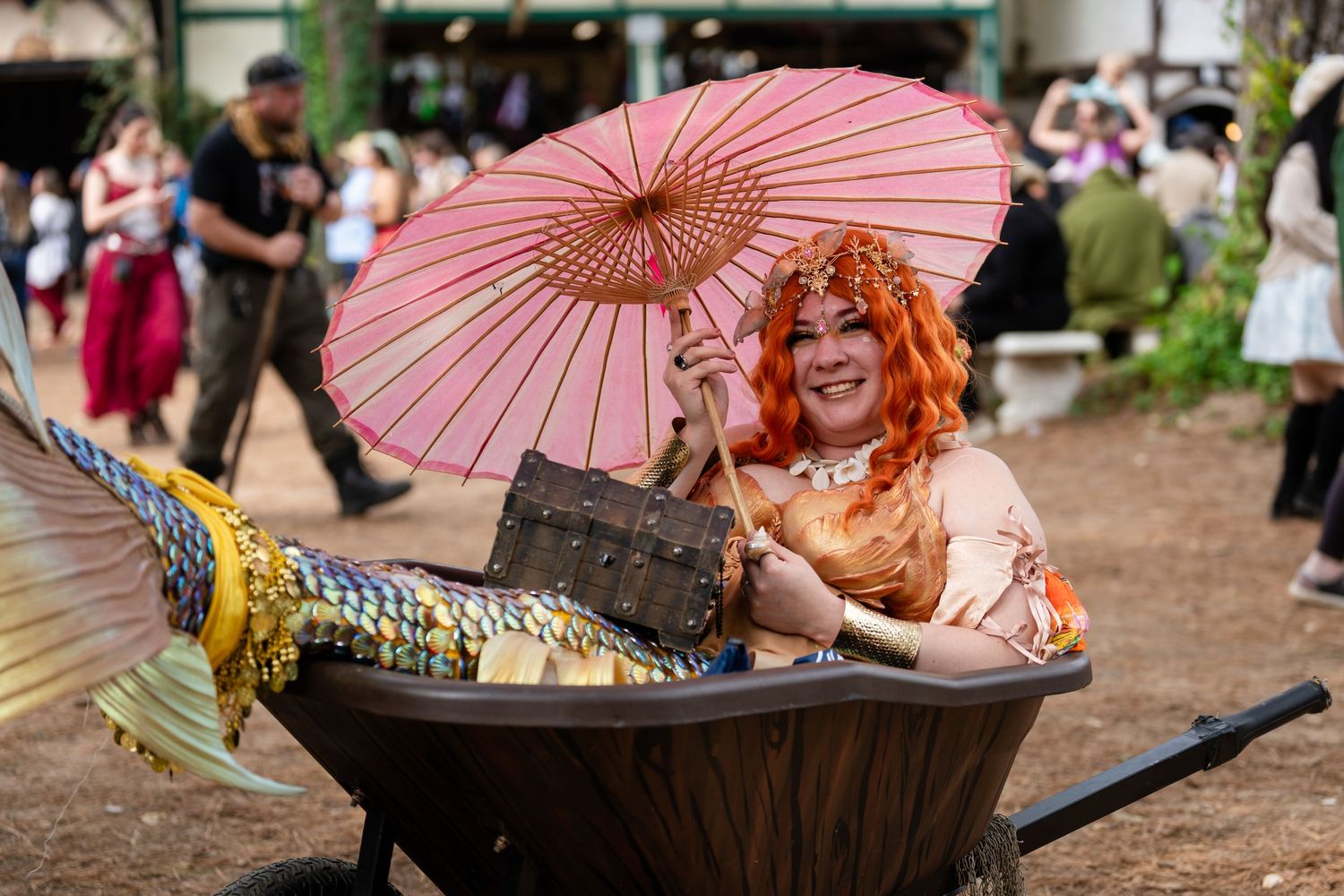 A whimsical performer in orange attire holds a pink parasol while reclining in a rustic wheelbarrow at a festival.