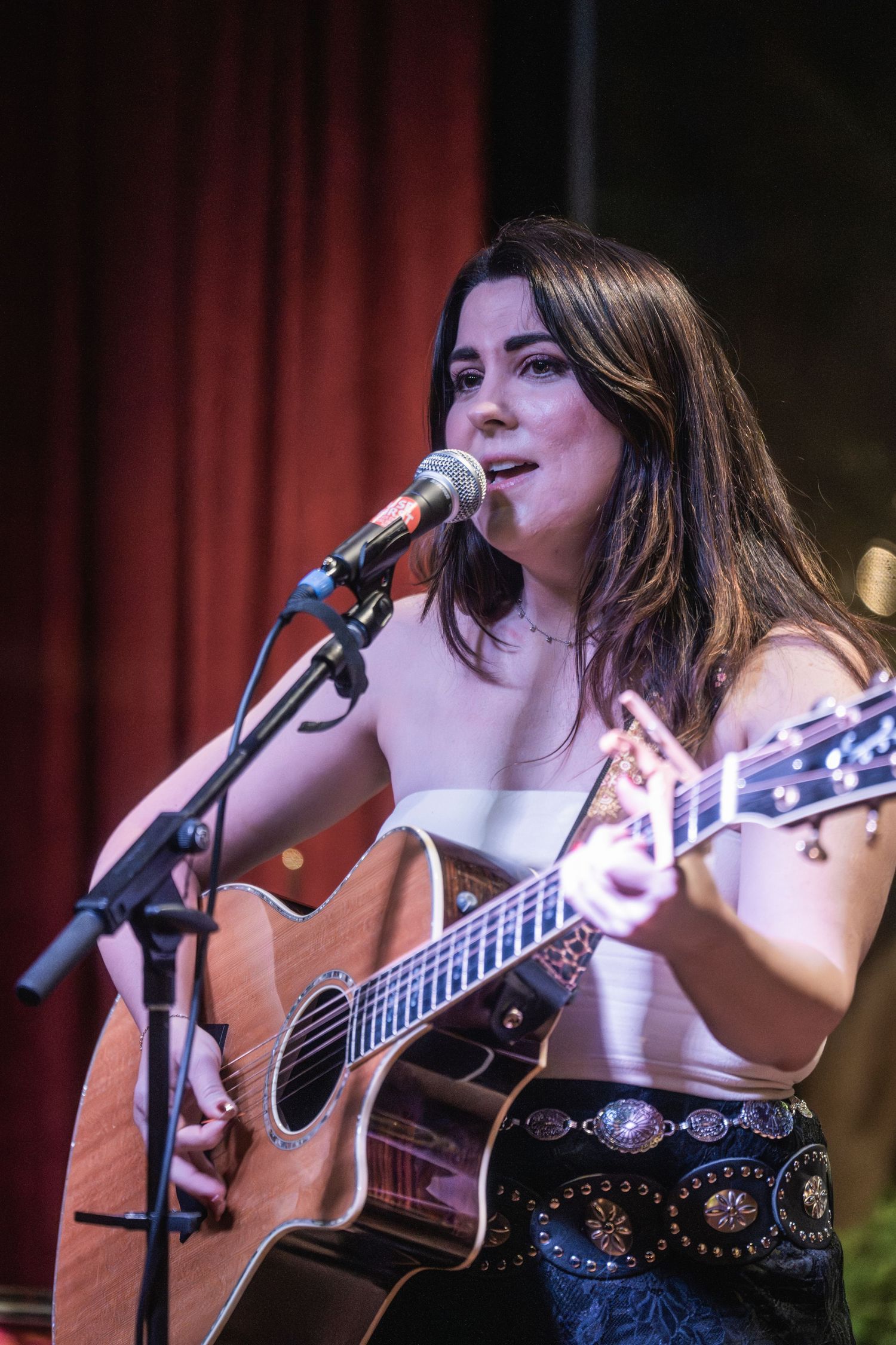 Musician performs on acoustic guitar in front of red curtain backdrop during live performance.