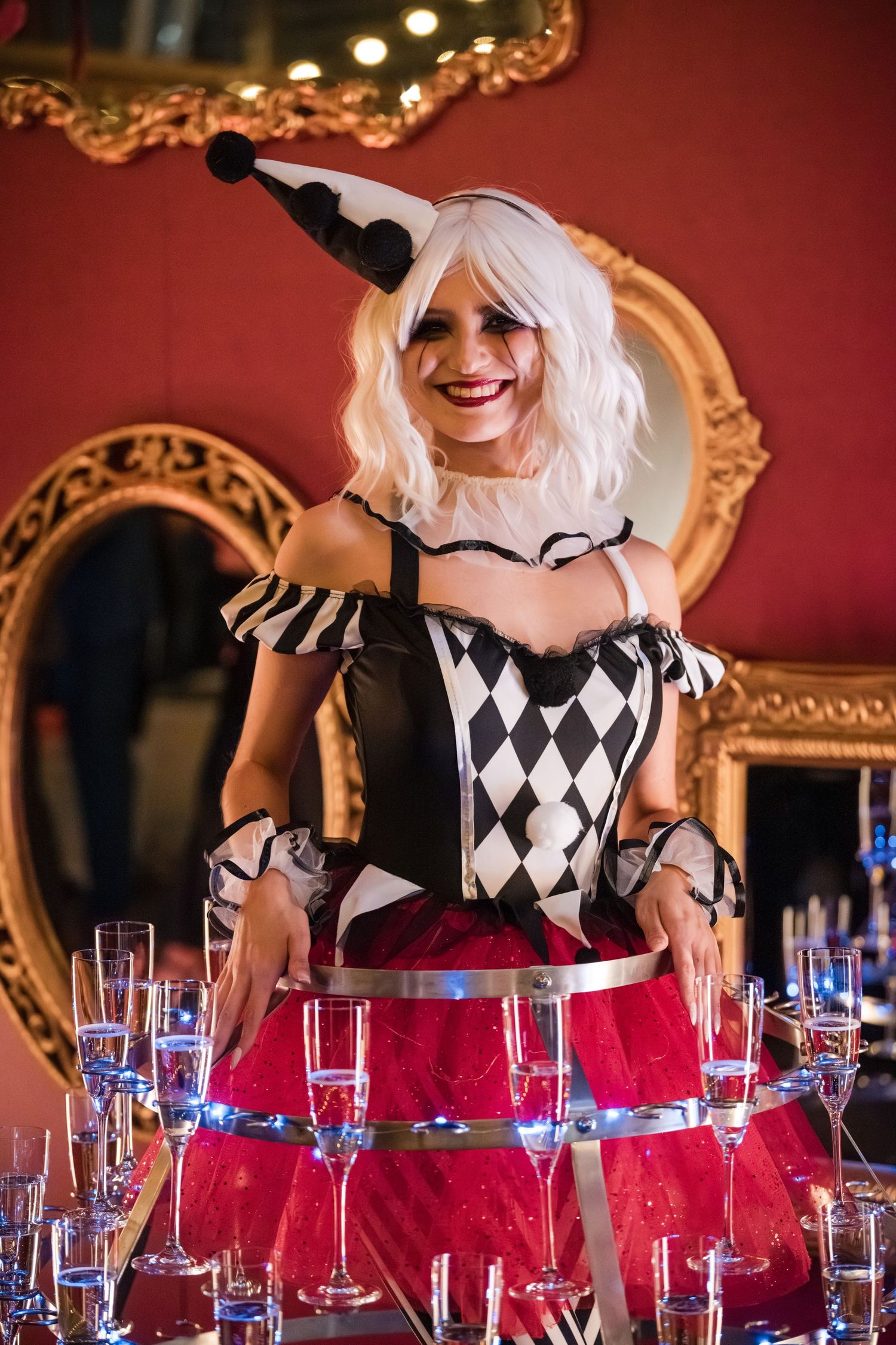 A person in a black and white diamond patterned dress arranges wine glasses on a table with a red tablecloth.