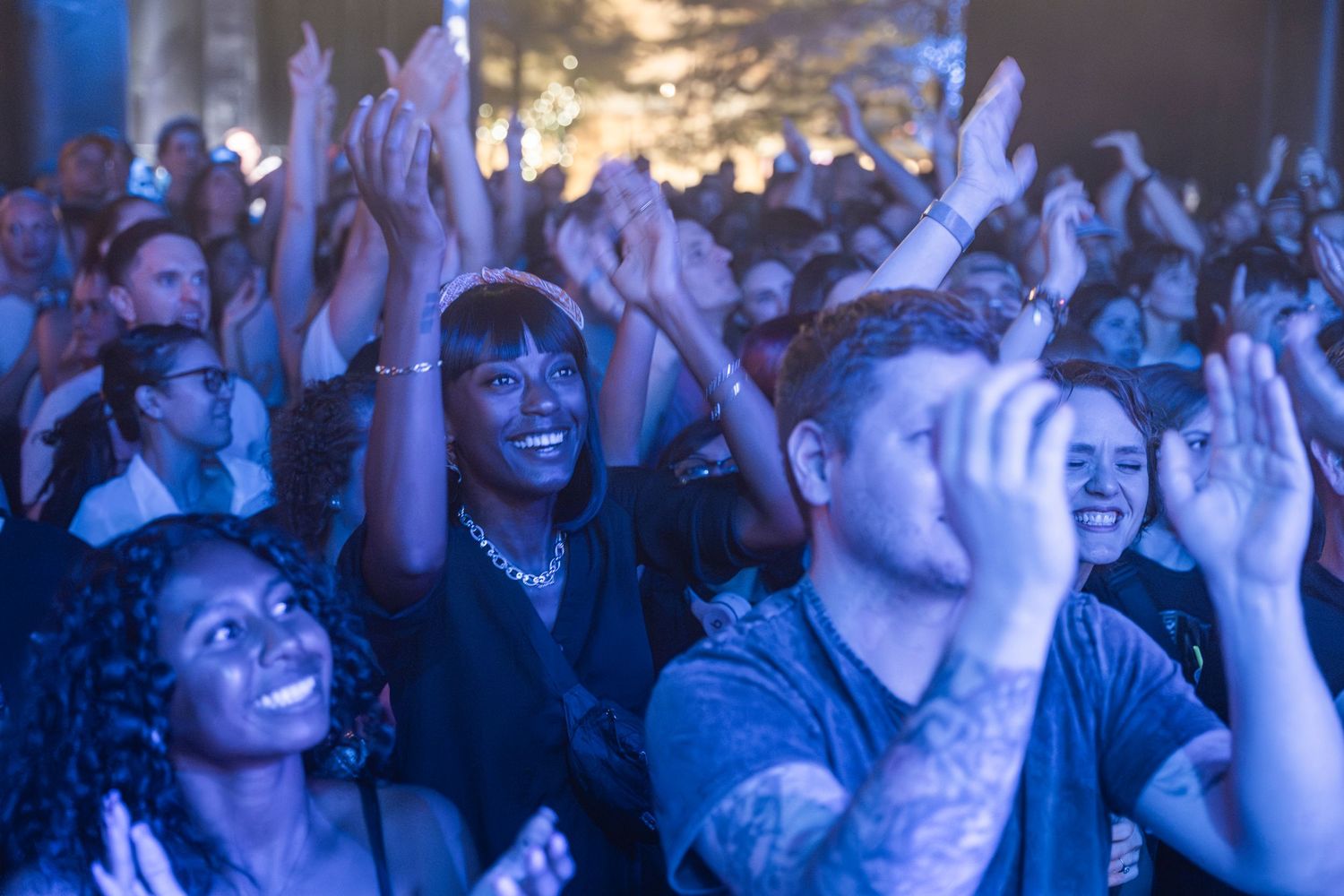 Concert crowd cheers and raises hands in excitement during a live music performance in a dark venue.