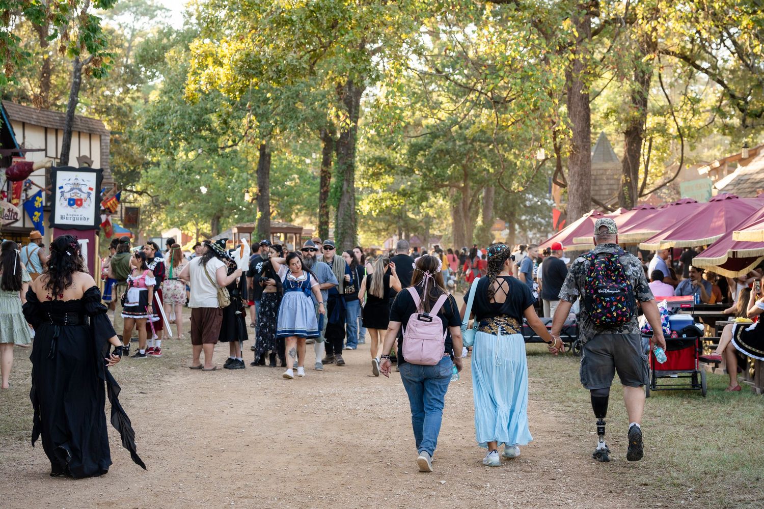 People gather in a park for a medieval festival with vendors and costumed attendees walking among trees.