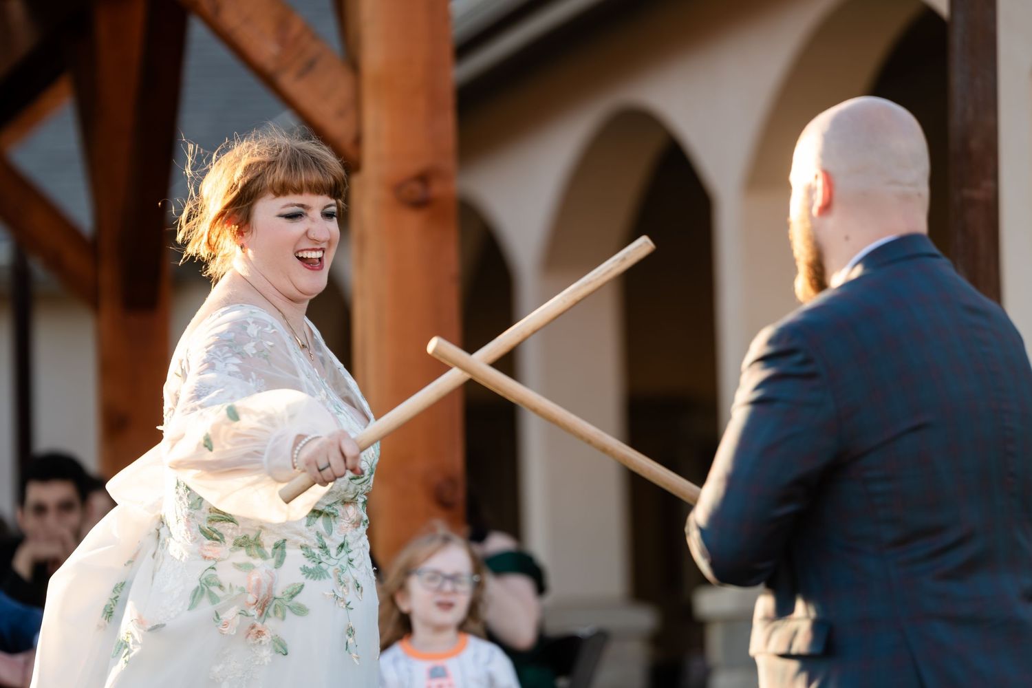 Two people in wedding attire playfully duel with drumsticks during a joyful outdoor celebration.