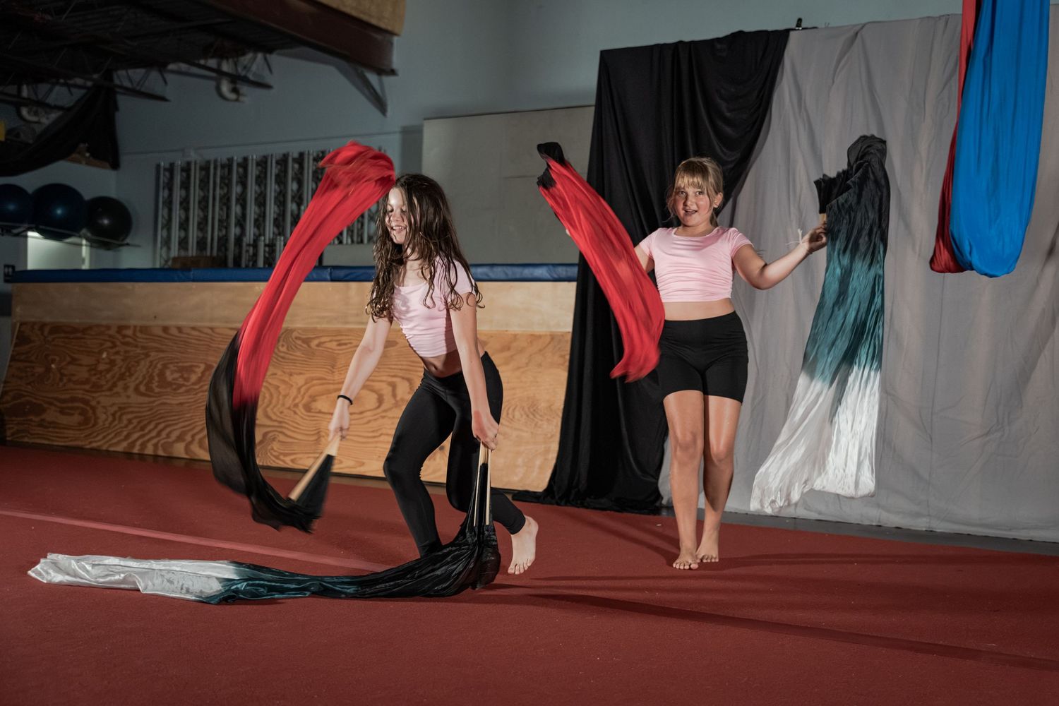 Two girls practice rhythmic gymnastics with flowing red and teal ribbons on a red mat in an indoor gym.
