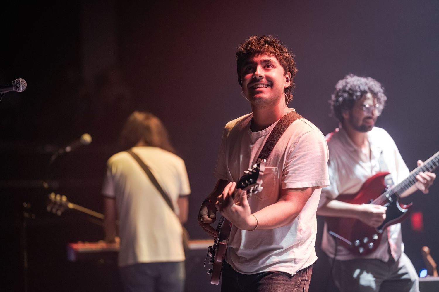 Smiling guitarist performs on stage with band members playing electric guitars in a live concert setting.