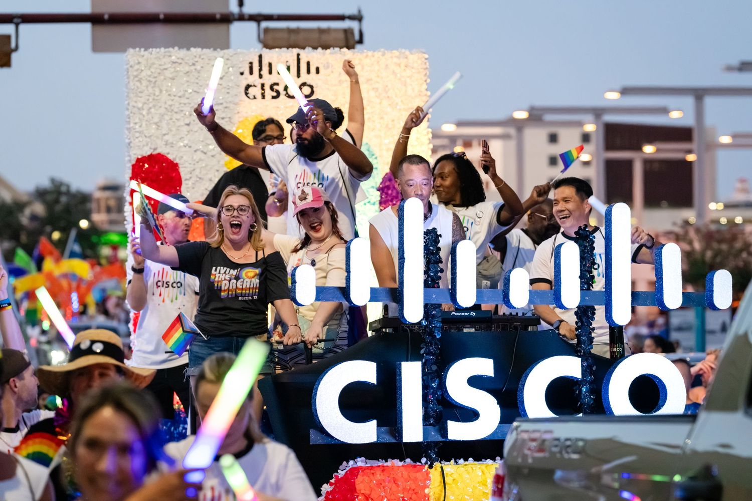 Cisco employees celebrate Pride on a colorful parade float, waving and cheering with rainbow flags and glow sticks.
