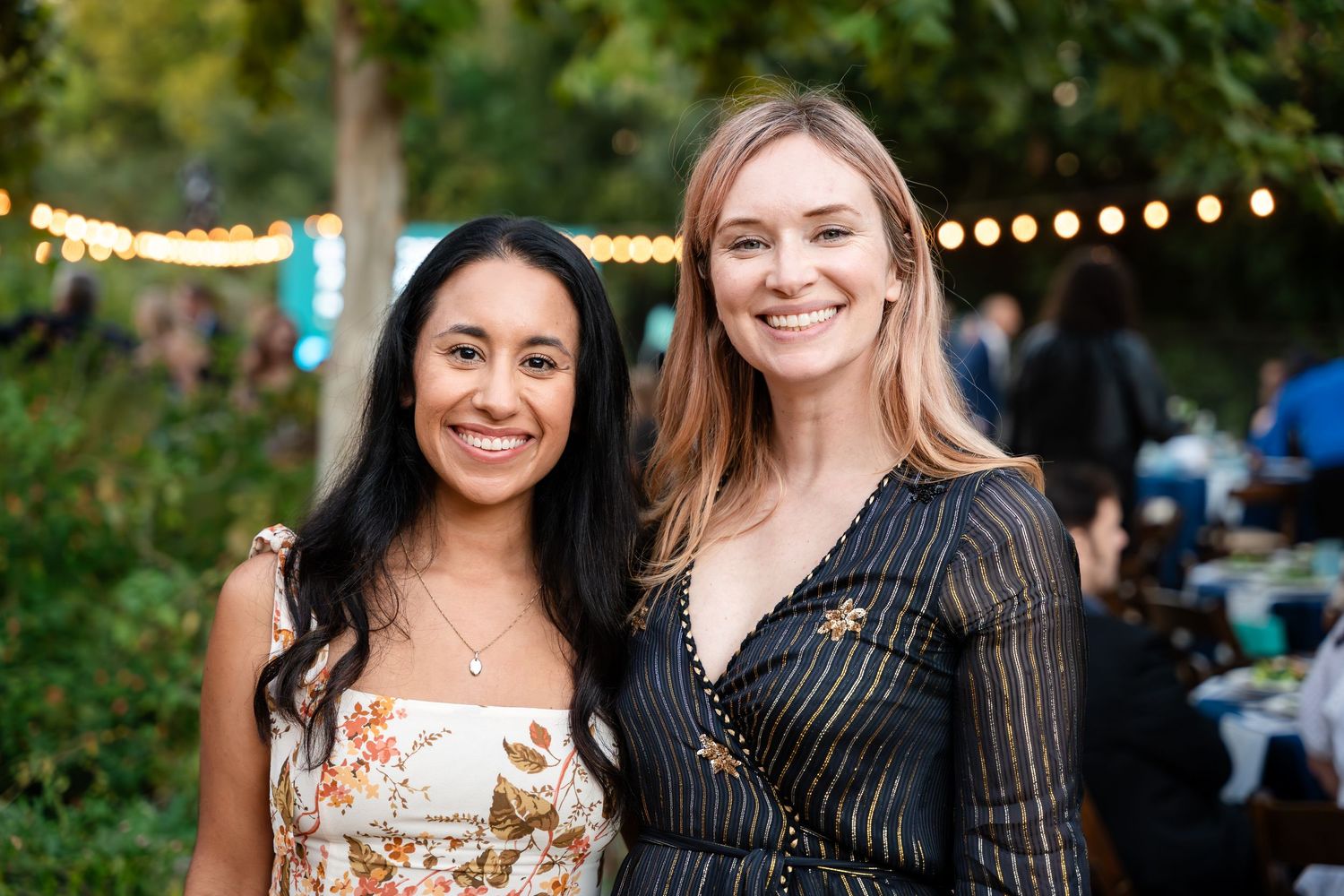Two people pose together outdoors at an evening event with string lights visible in the background.