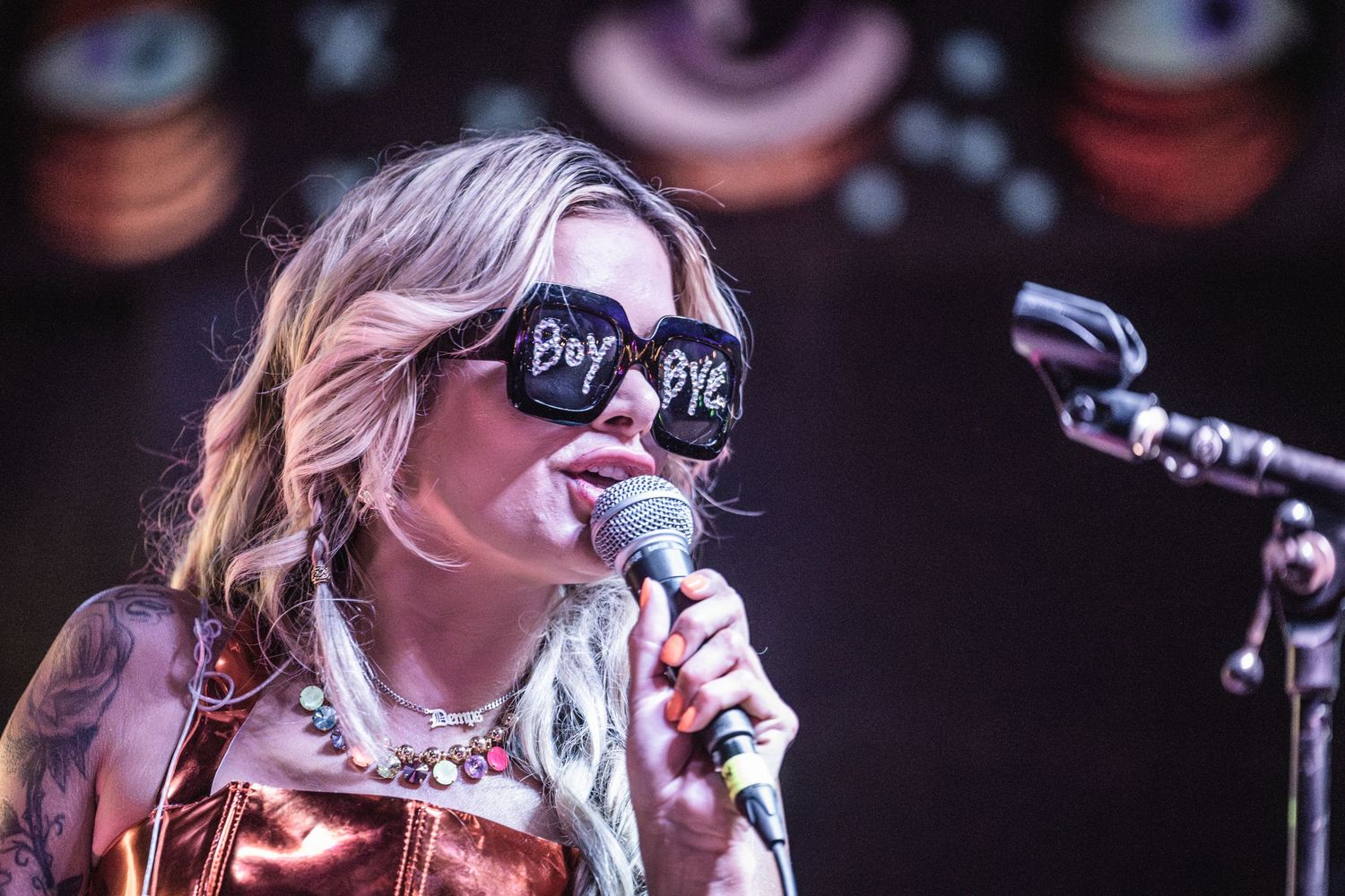 A performer in sunglasses sings passionately into a microphone on a dark stage with dramatic lighting.