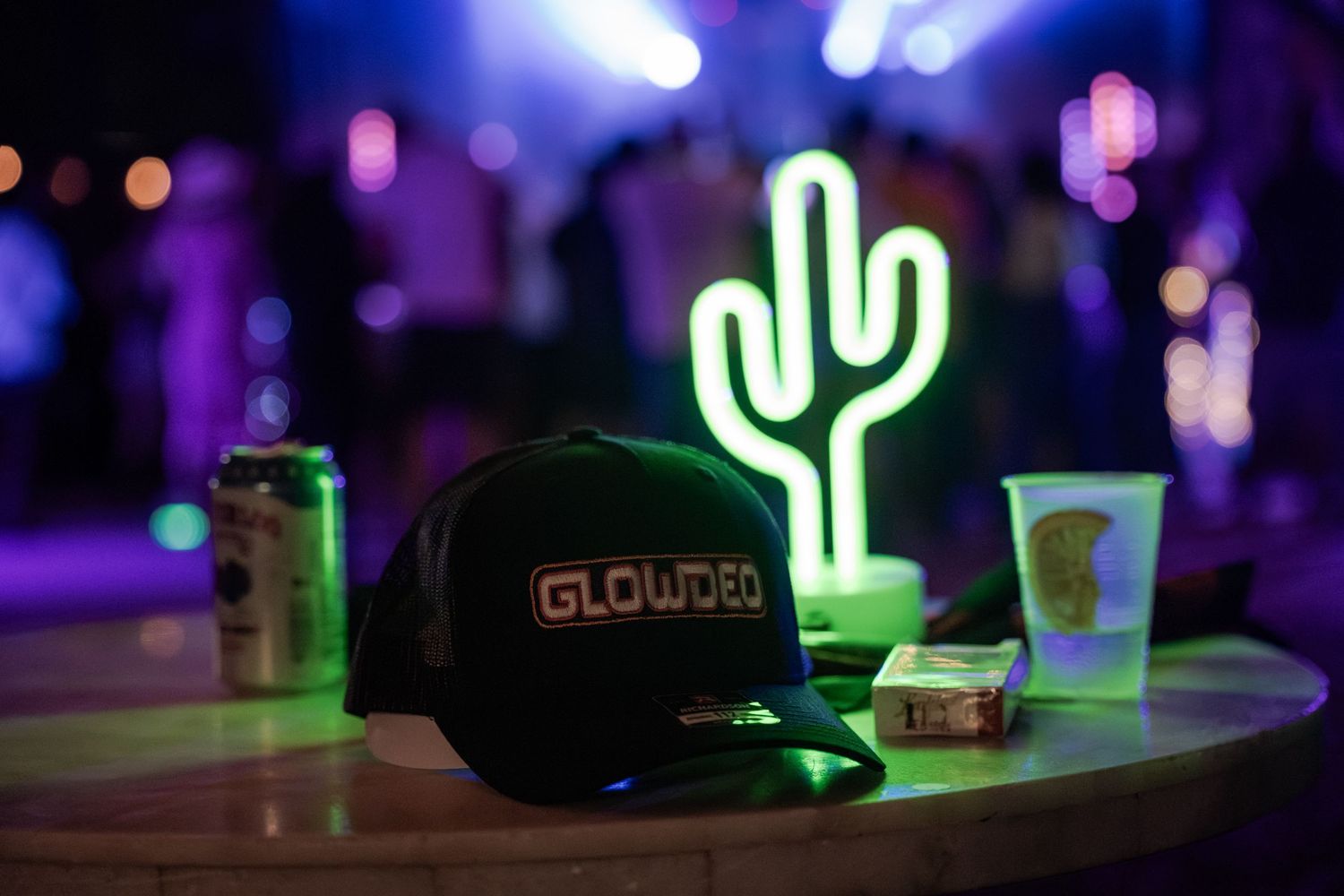 Glowded snapback hat beside a neon cactus sign on a table at a vibrant nightclub event.