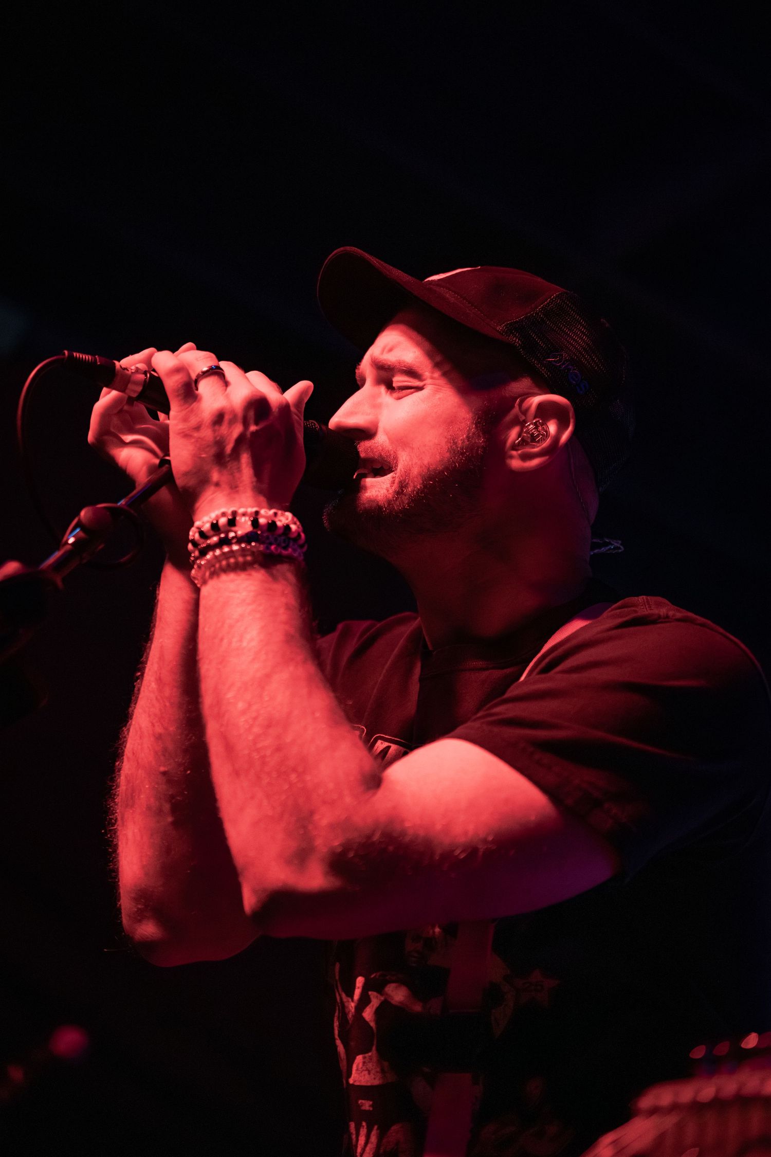 Male singer performing live on stage under red lights, holding microphone with both hands, wearing cap and bracelets.