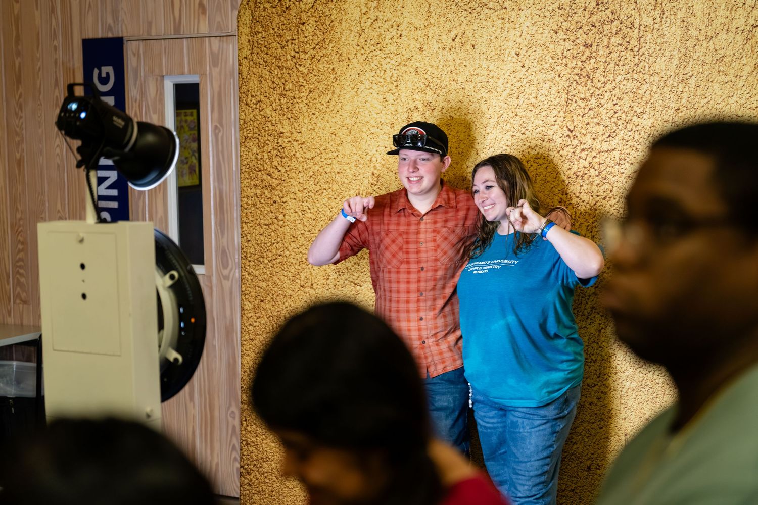 Two people pose flexing their arms and smiling against a textured gold wall indoors.