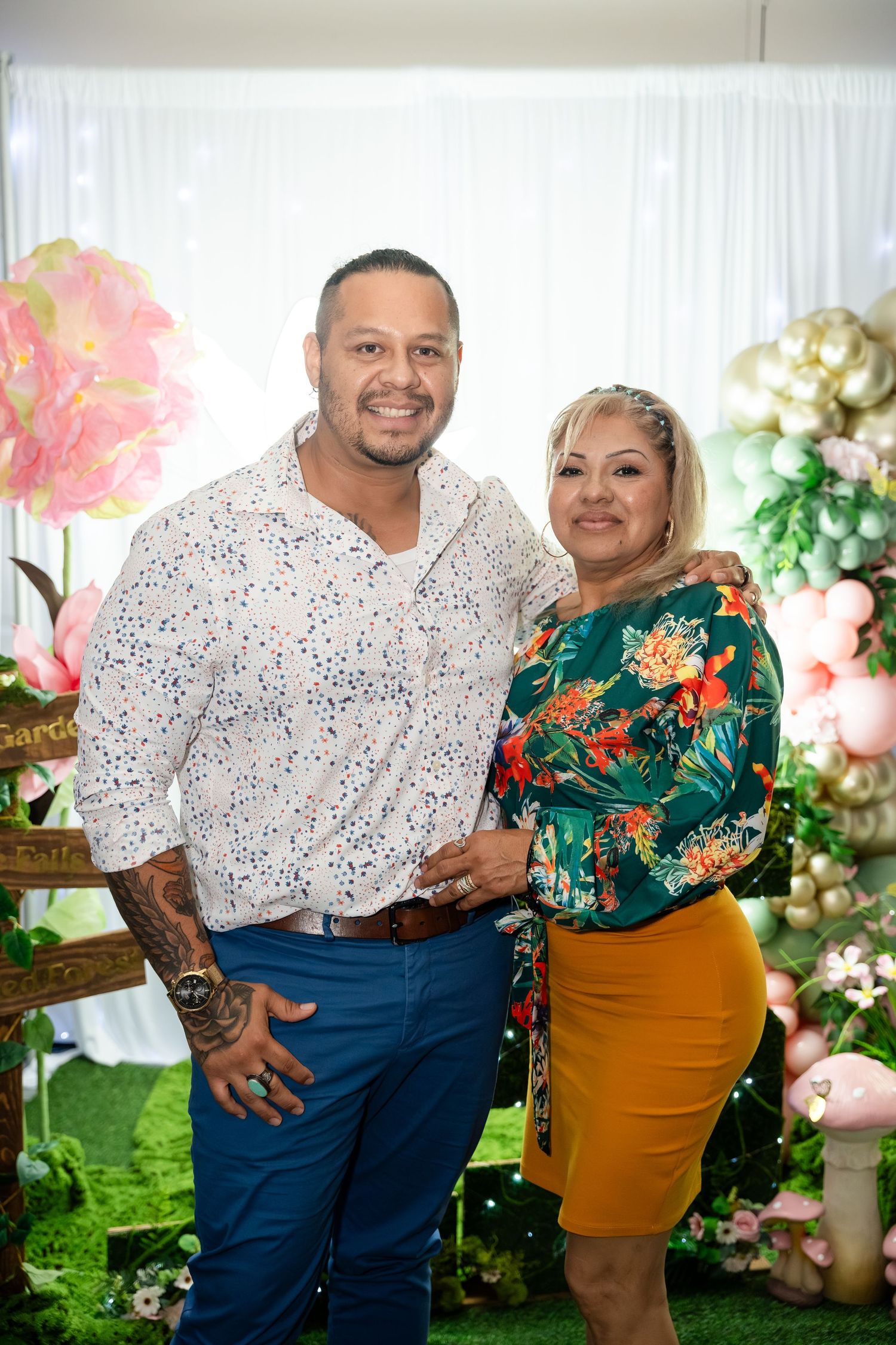 A man and woman pose together smiling at a festive garden party with balloon decorations and floral backdrop.