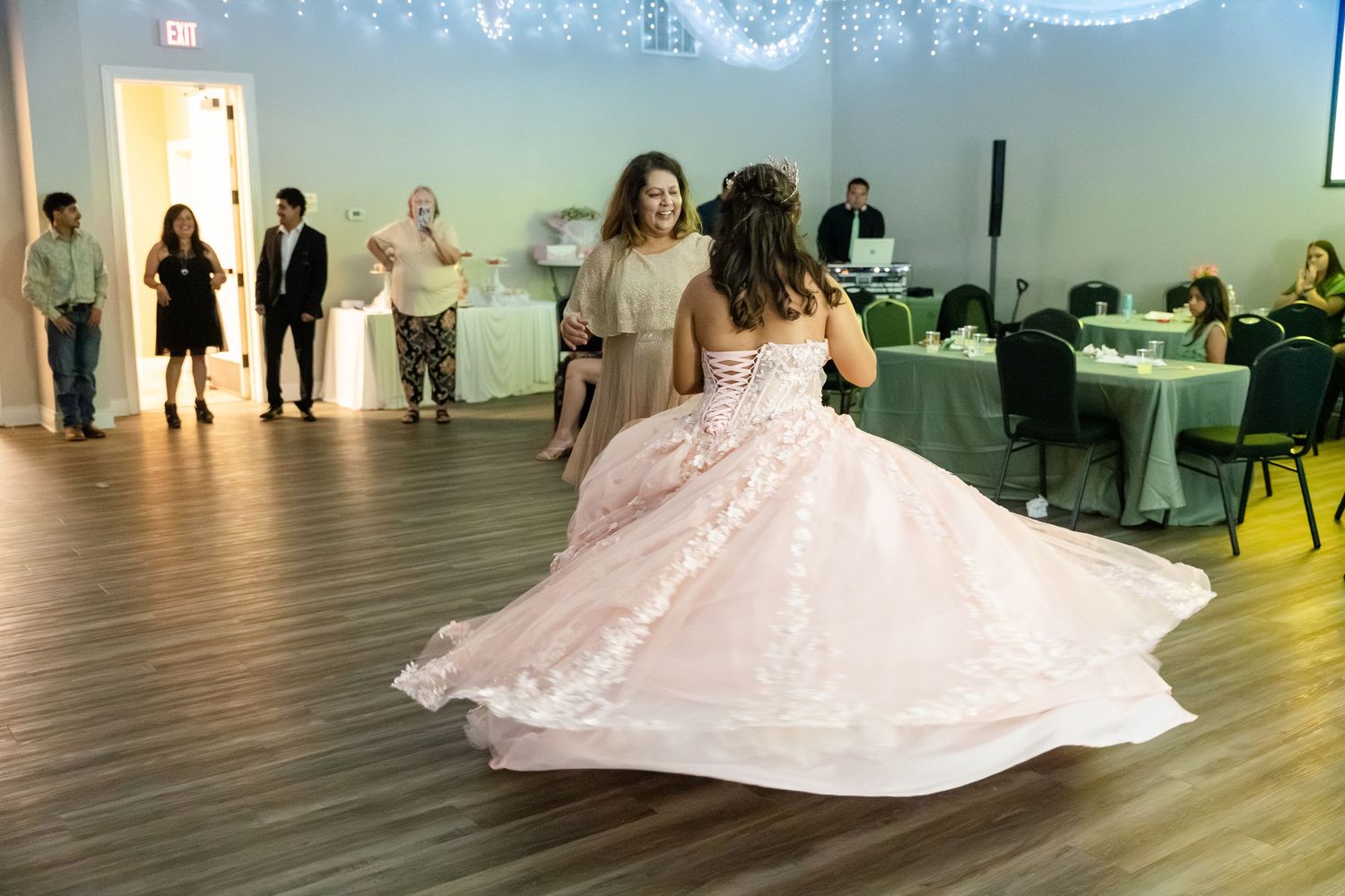 Two women dancing gracefully at a quinceañera celebration, one in an elegant pink ball gown, in a decorated event hall.