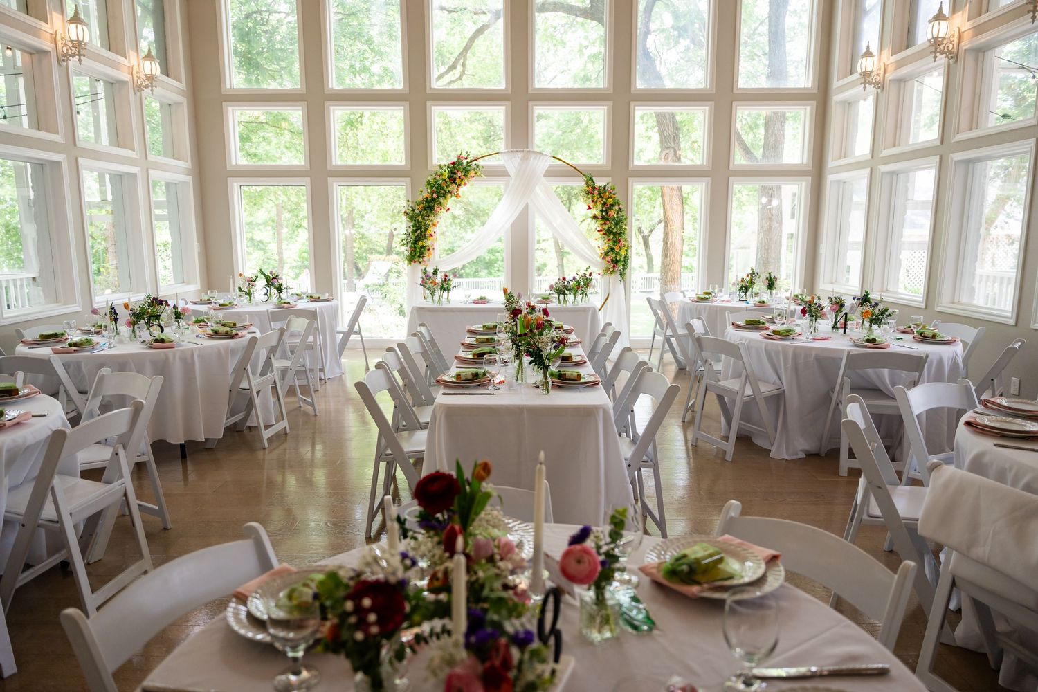 Elegant wedding reception hall with white round tables, floral centerpieces, and a greenery arch framed by large windows.