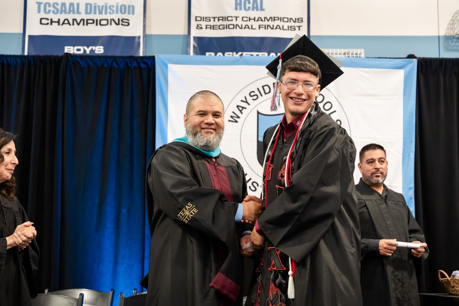 Two graduates in caps and gowns shake hands on stage at a Wayside School graduation ceremony.