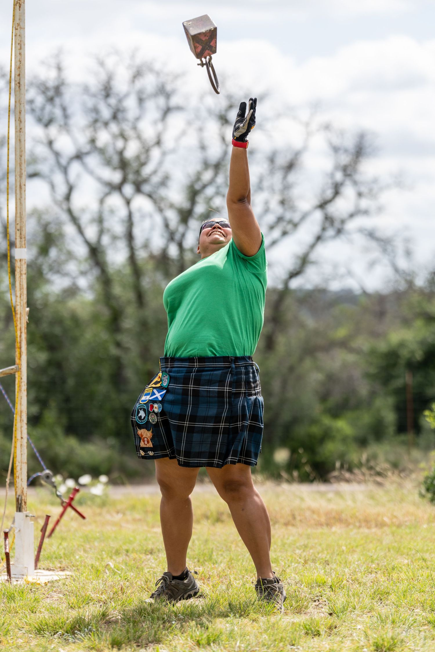 Athlete in green shirt and tartan kilt tosses weight overhead at Highland Games competition outdoors.