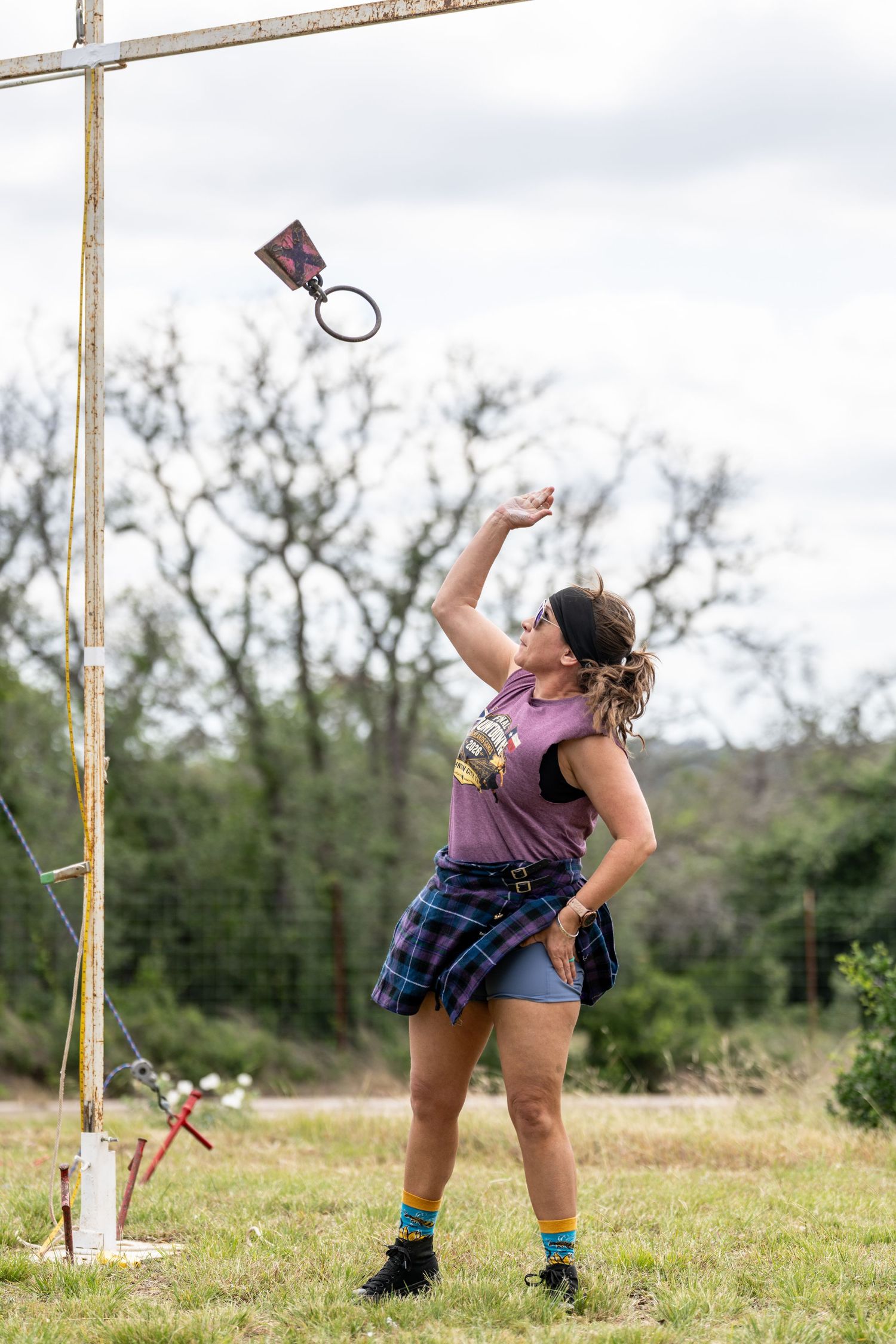 Woman in plaid skirt and purple tank top tosses weight over bar at Highland Games competition outdoors.