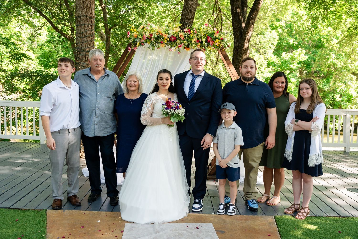 Wedding party poses outdoors on wooden deck surrounded by lush green trees with floral arch backdrop.