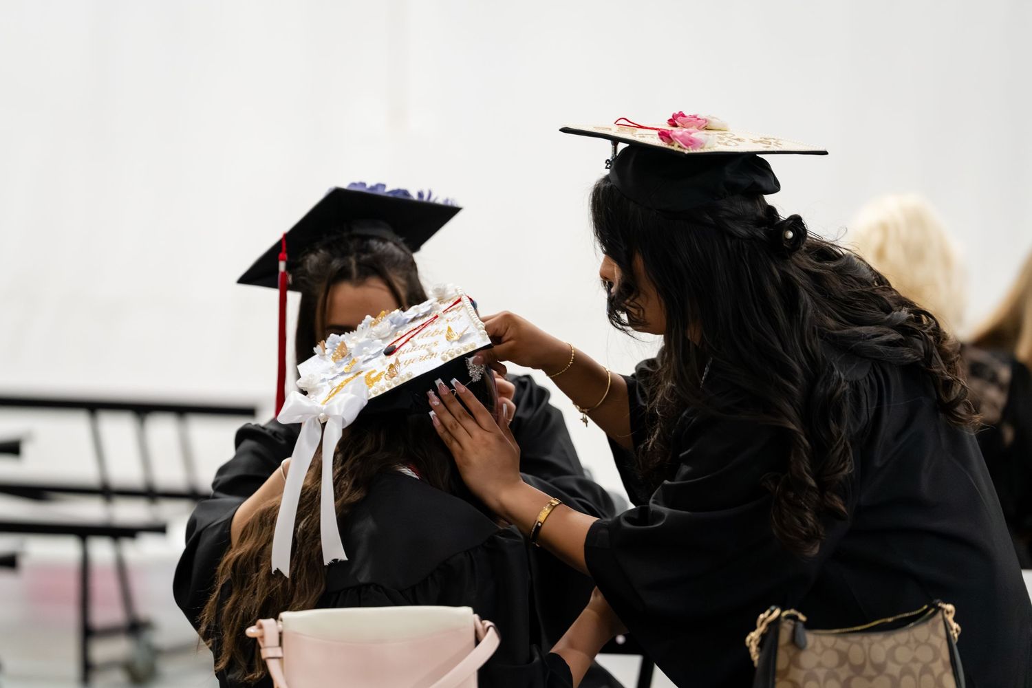 Two graduates in black robes and decorated caps at a graduation ceremony, one adjusting the other's cap.