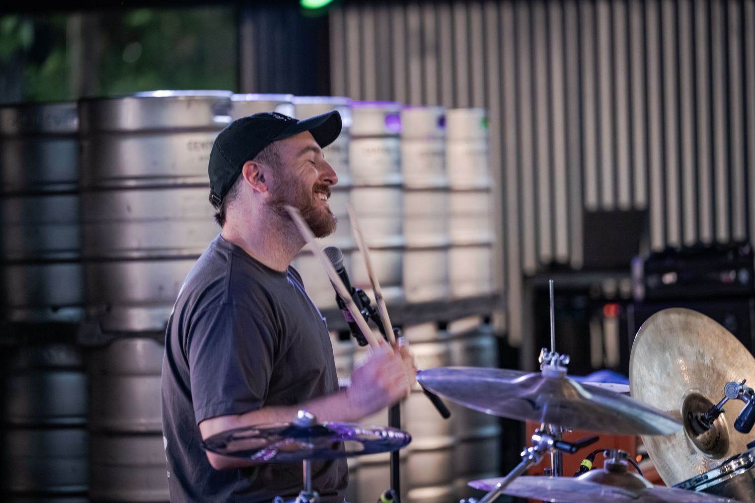 Smiling male drummer playing cymbals and drums at an indoor venue with metal kegs in the background.