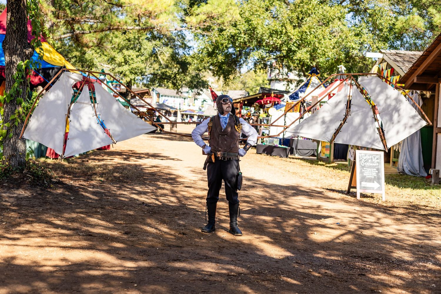 Colorful flags and banners decorate a shaded outdoor festival area where people gather on a sunny day.