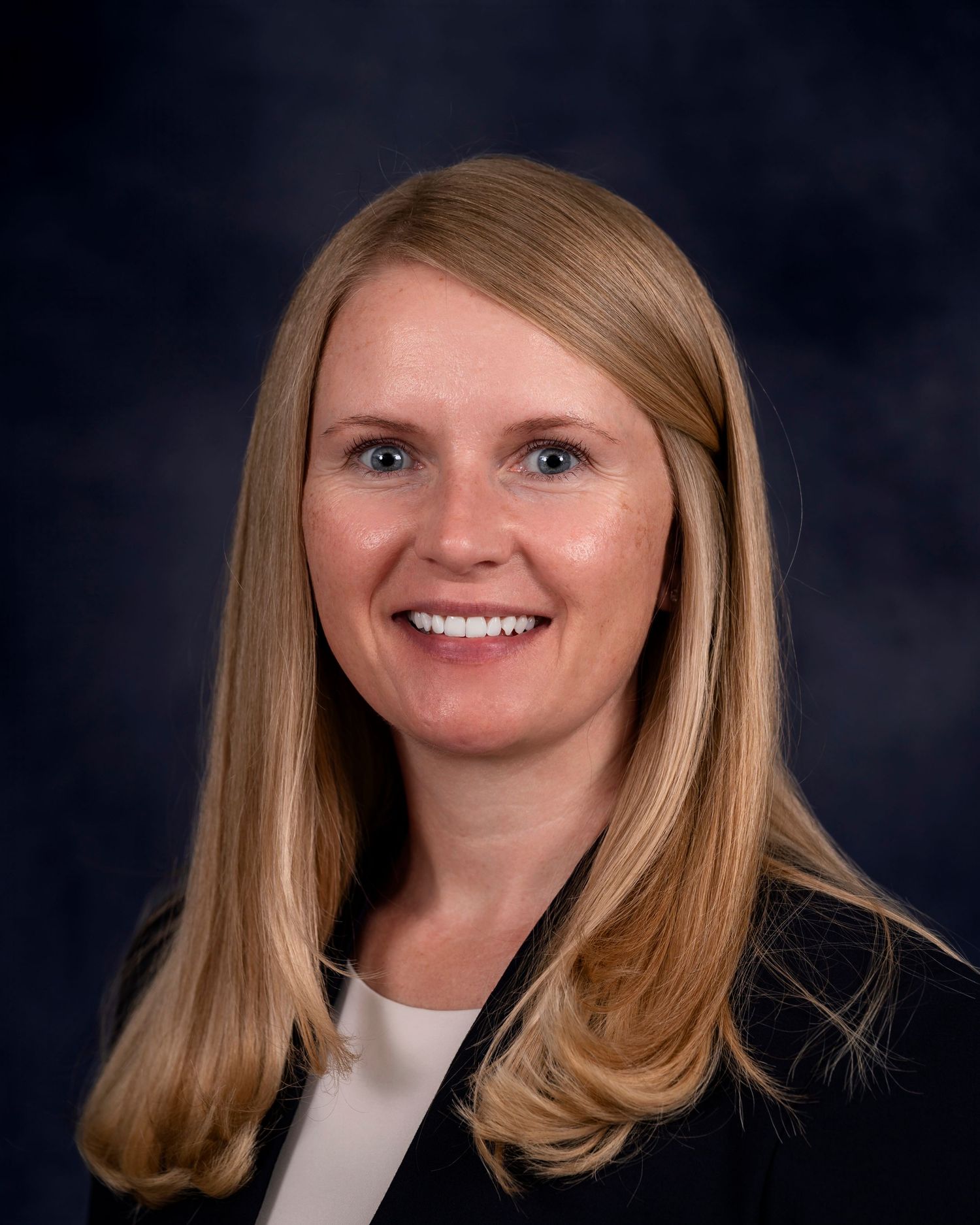 Professional headshot portrait against dark background with blonde hair and white top.