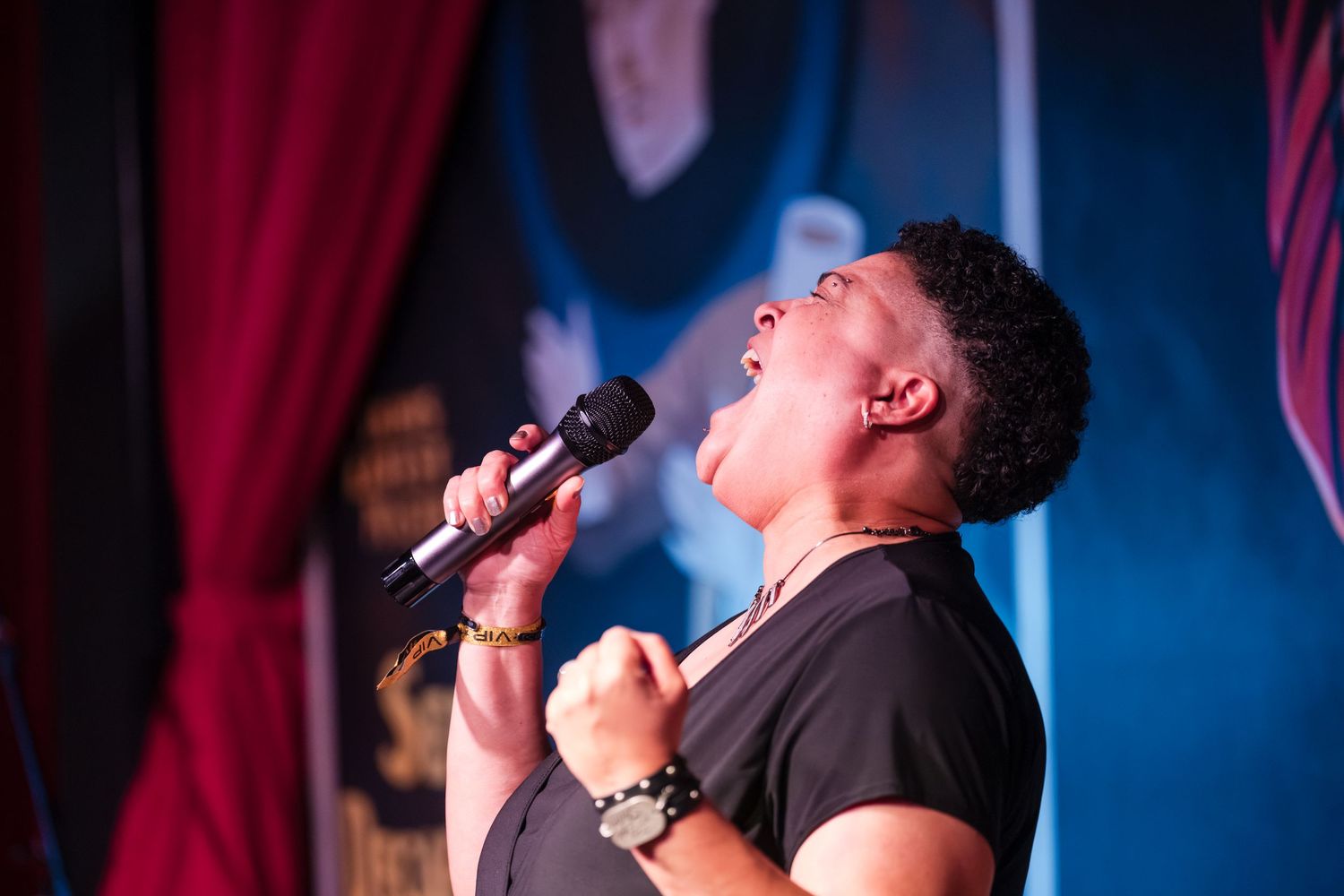 A passionate performer sings into a microphone on stage with dramatic red and blue lighting in the background.