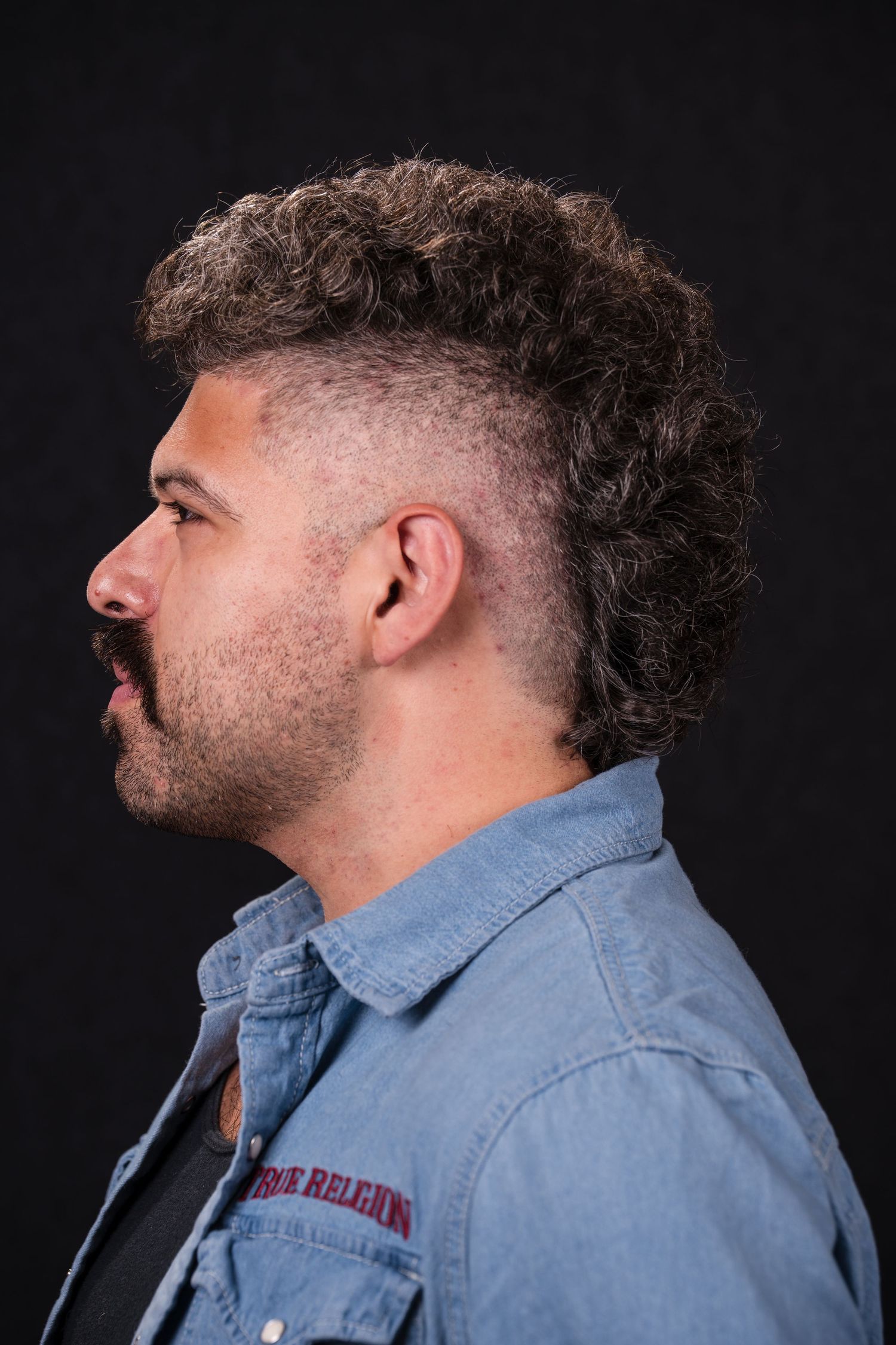 Side profile portrait of person with curly dark hair and faded sides wearing denim jacket against black background.