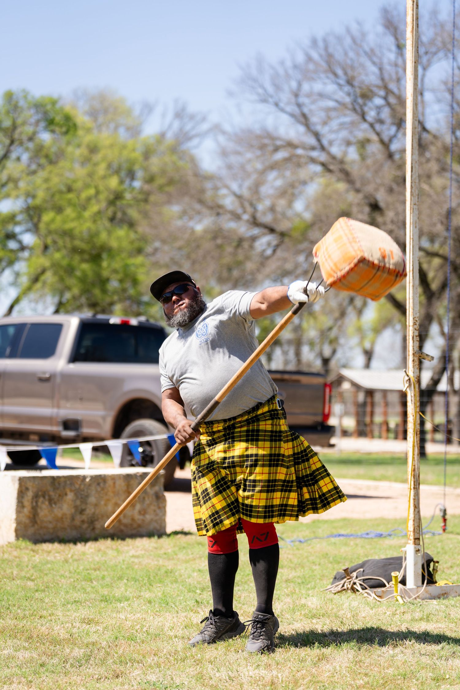 Man in yellow tartan kilt performing Scottish Highland Games hammer throw outdoors on a sunny day.