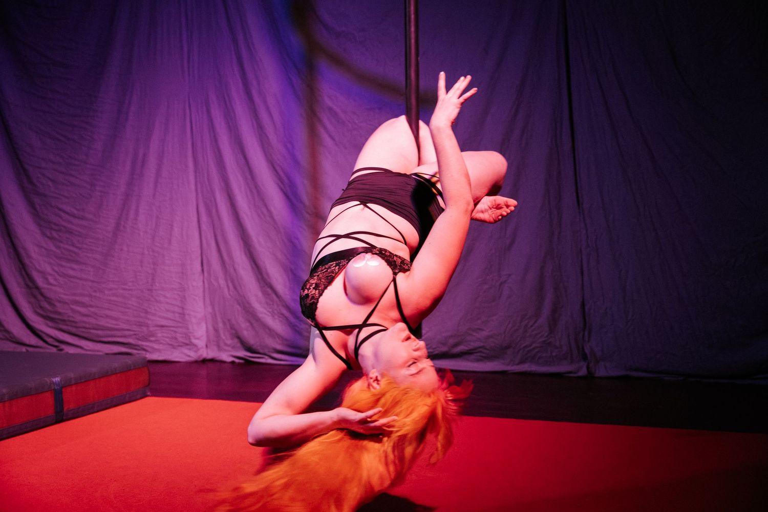 Woman performing an acrobatic pole dance routine on a red stage with purple draped backdrop.