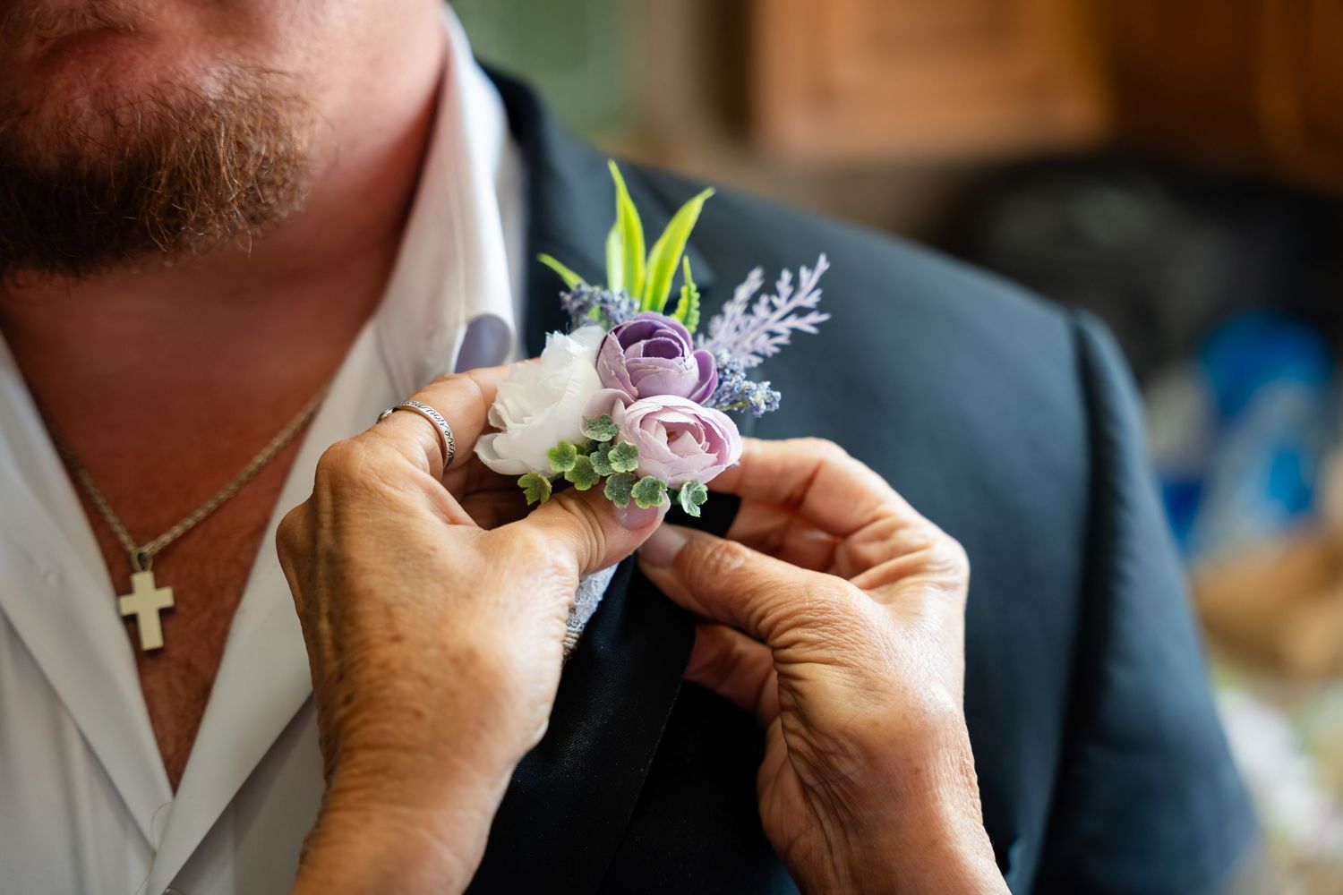 Hands pinning purple and white floral boutonniere on groom's navy suit lapel at wedding.