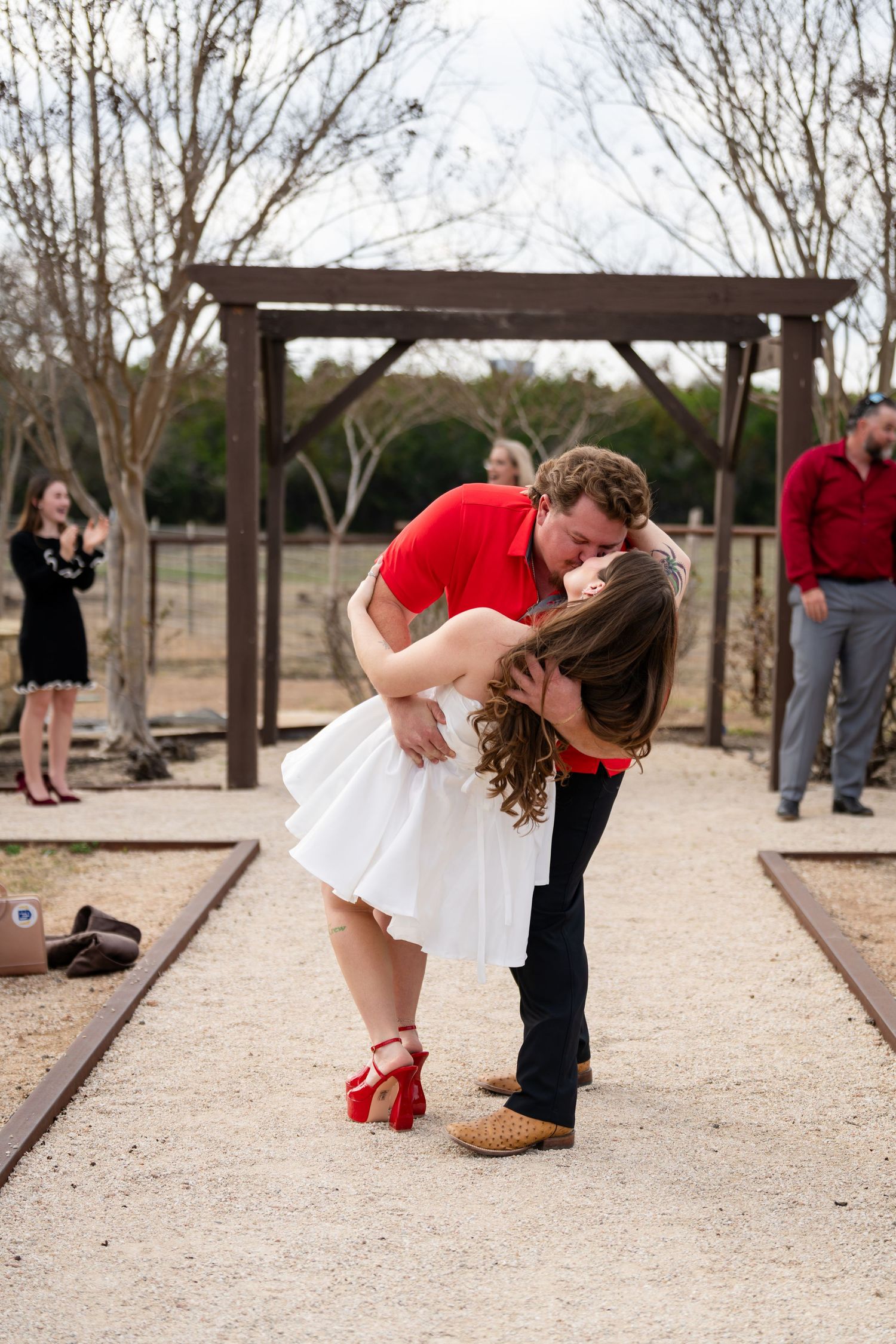 Couple sharing a romantic dip kiss at outdoor wedding ceremony, bride in white dress and red heels, groom in red polo shirt.