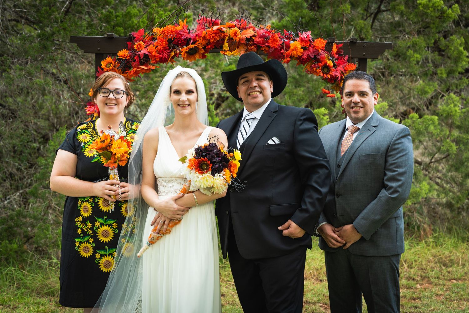 Wedding group portrait under a vibrant floral arch with sunflower bouquets.