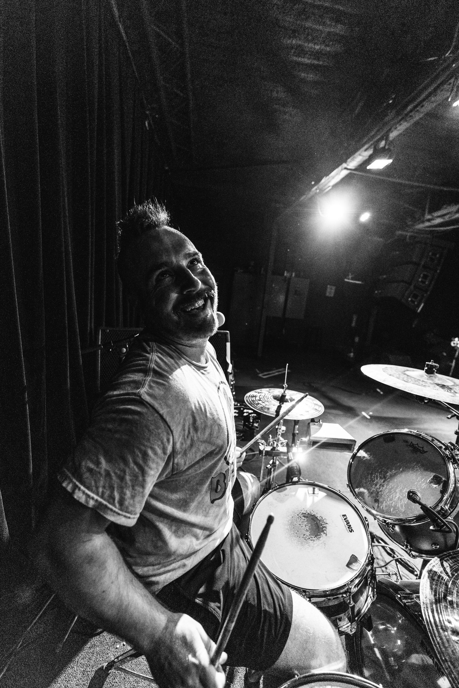 Drummer performs enthusiastically behind drum kit in dramatic black and white lighting.
