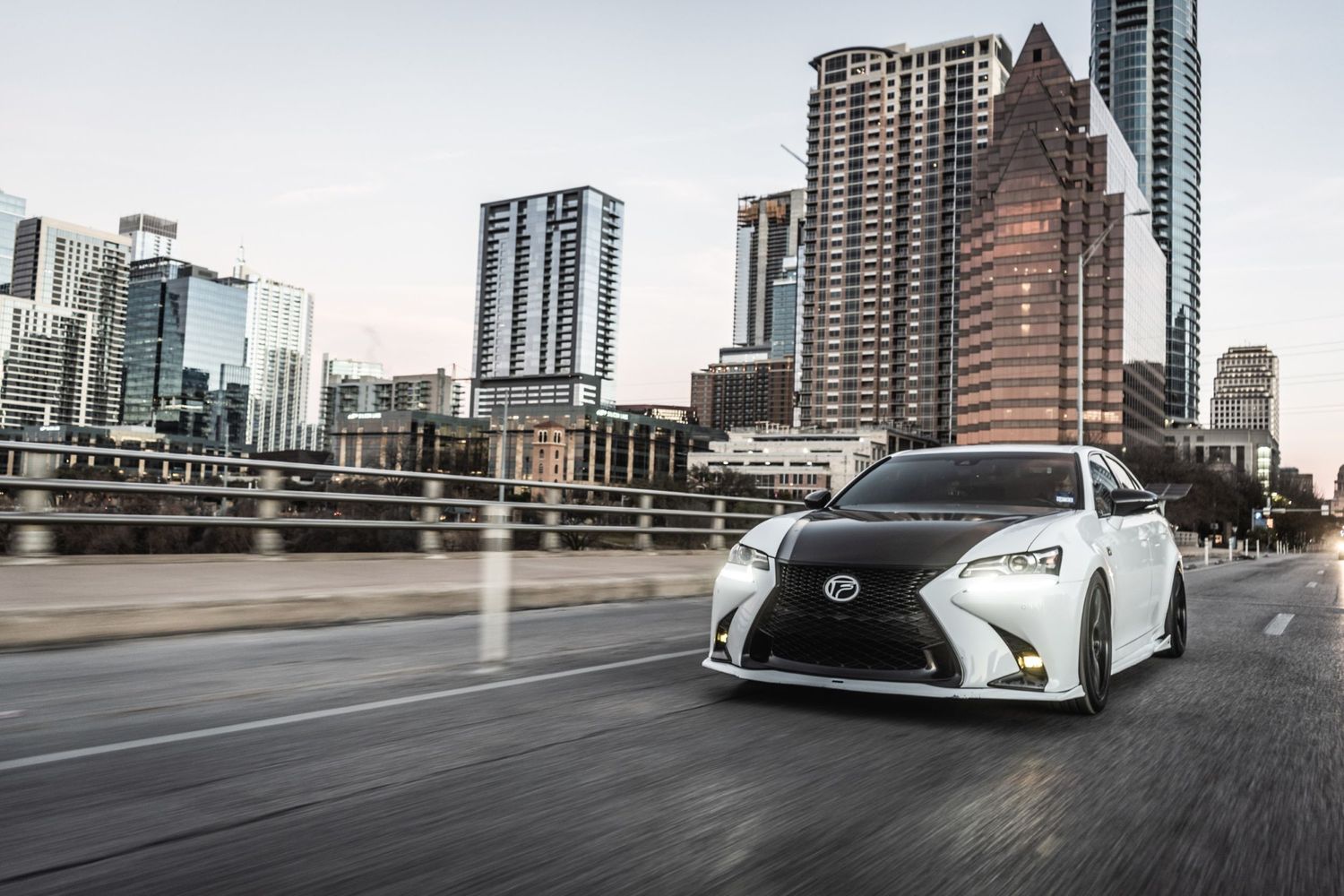 Lowered white Lexus cruising past modern Austin skyline on wet streets.