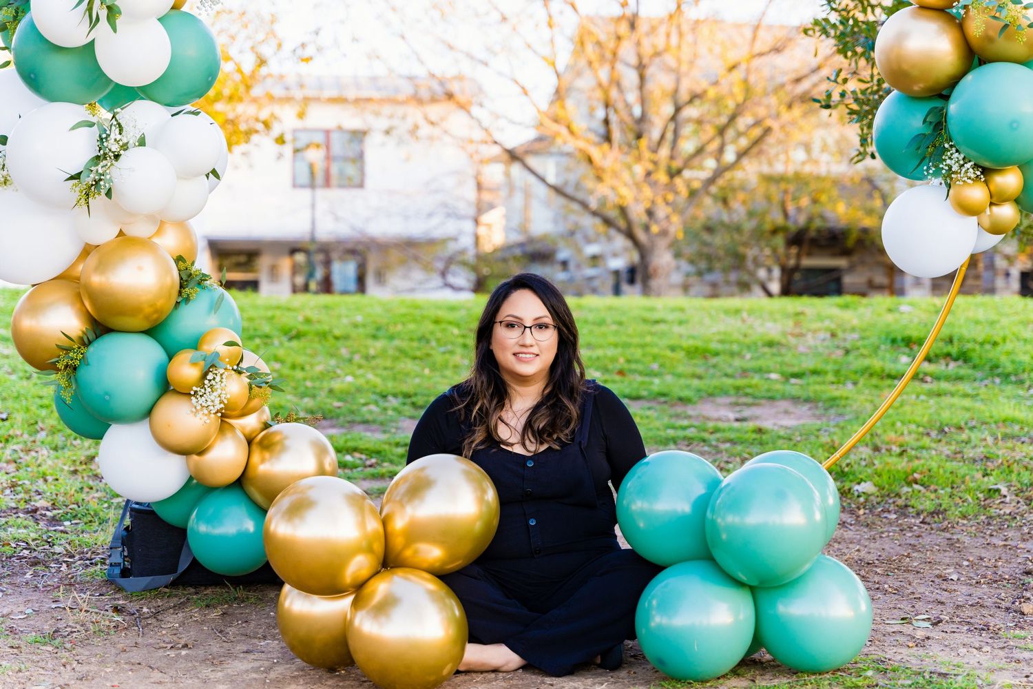 Person sitting beside luxurious balloon arch with gold and teal colors in outdoor setting.