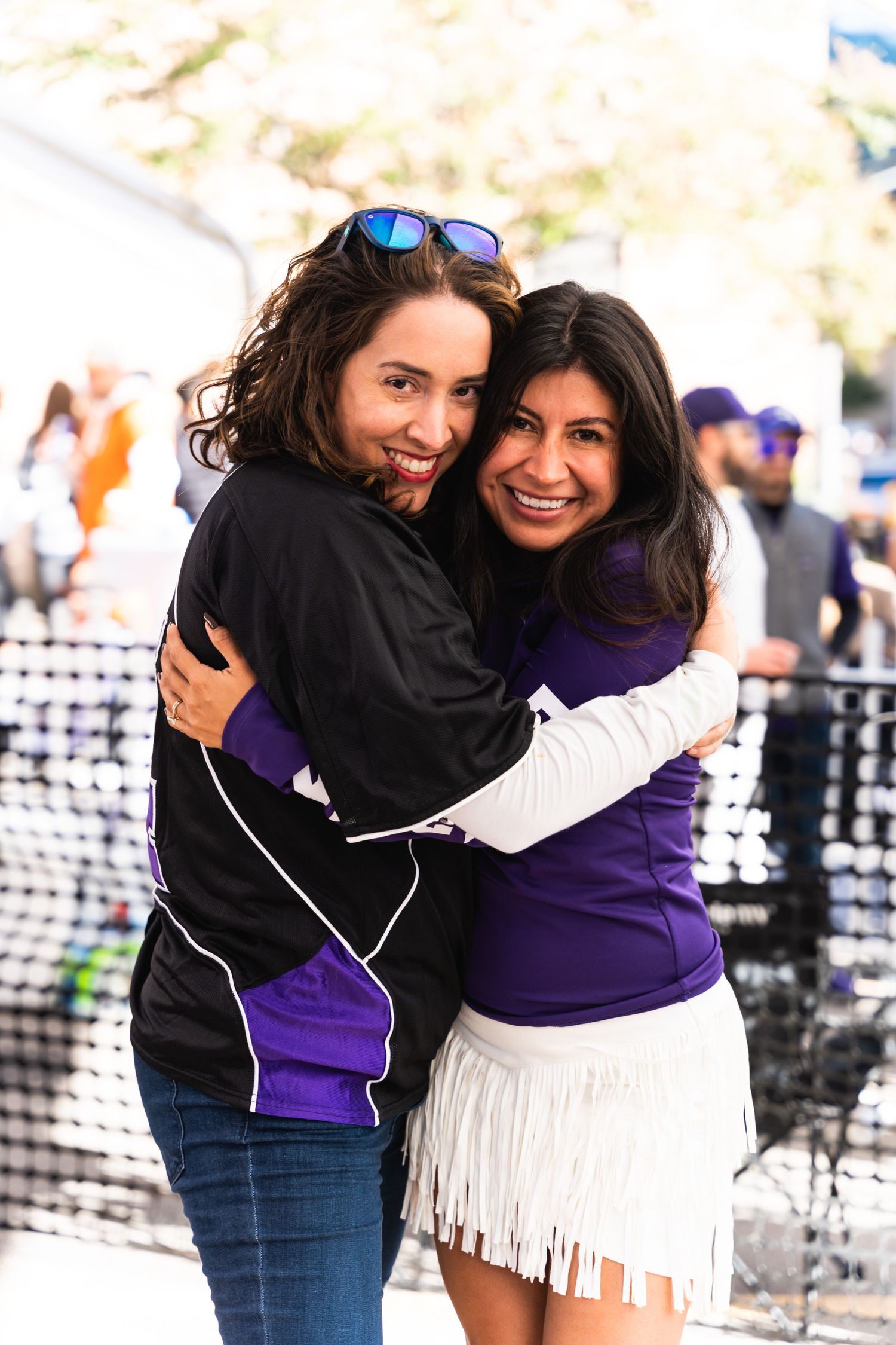 Two friends embracing and smiling at an outdoor festival with event barriers visible behind them.