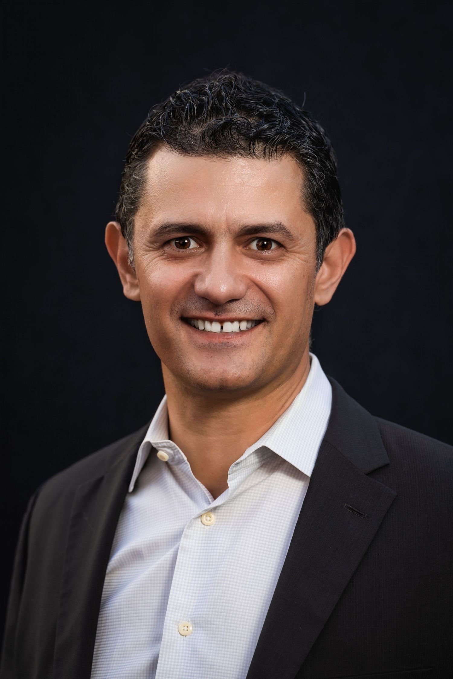 Professional headshot of man in dark suit jacket and white shirt against black background.